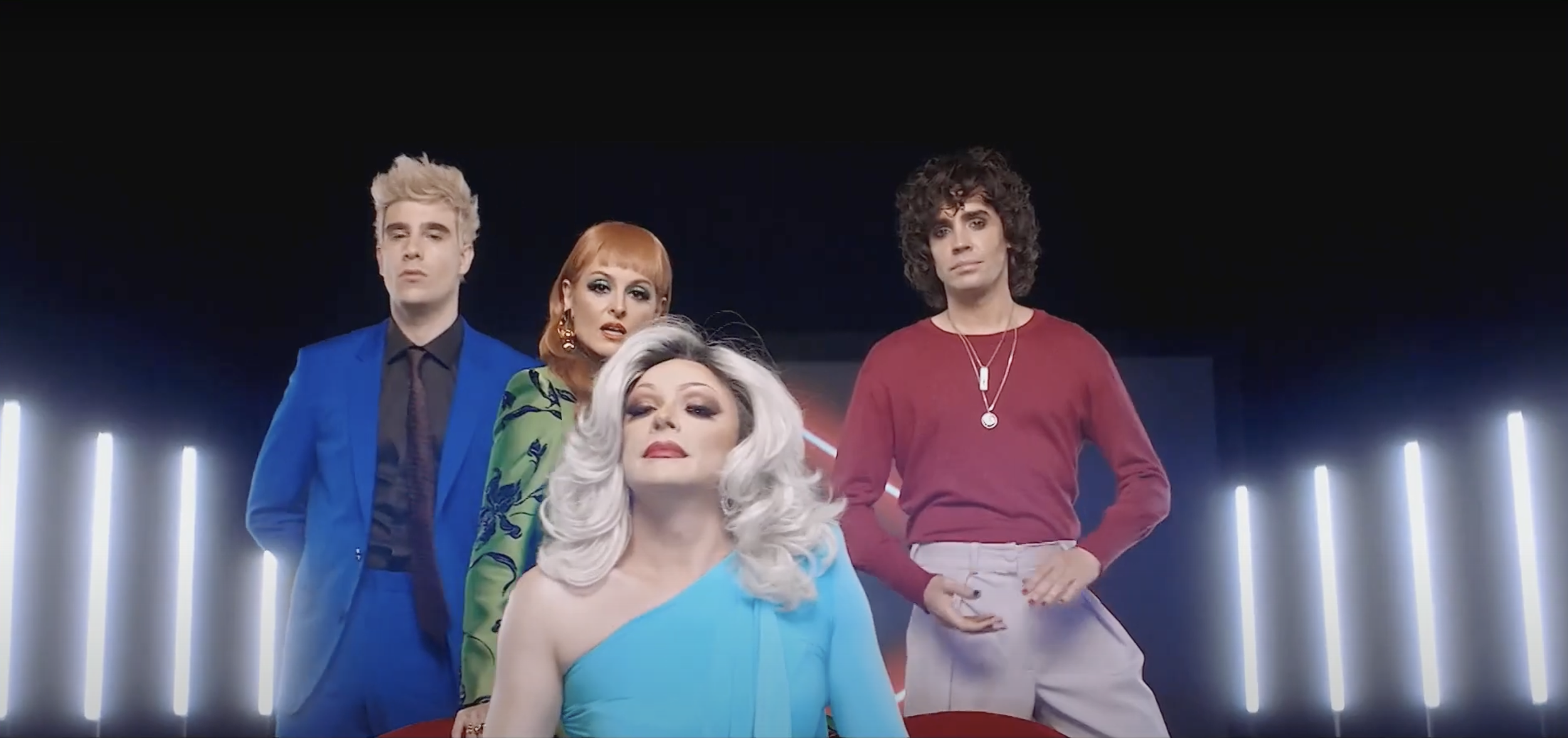 Four people standing with neon lights in the background, one in a bright blue suit, one in a patterned green dress, one in a red shirt, and one in a light blue one-shoulder gown.