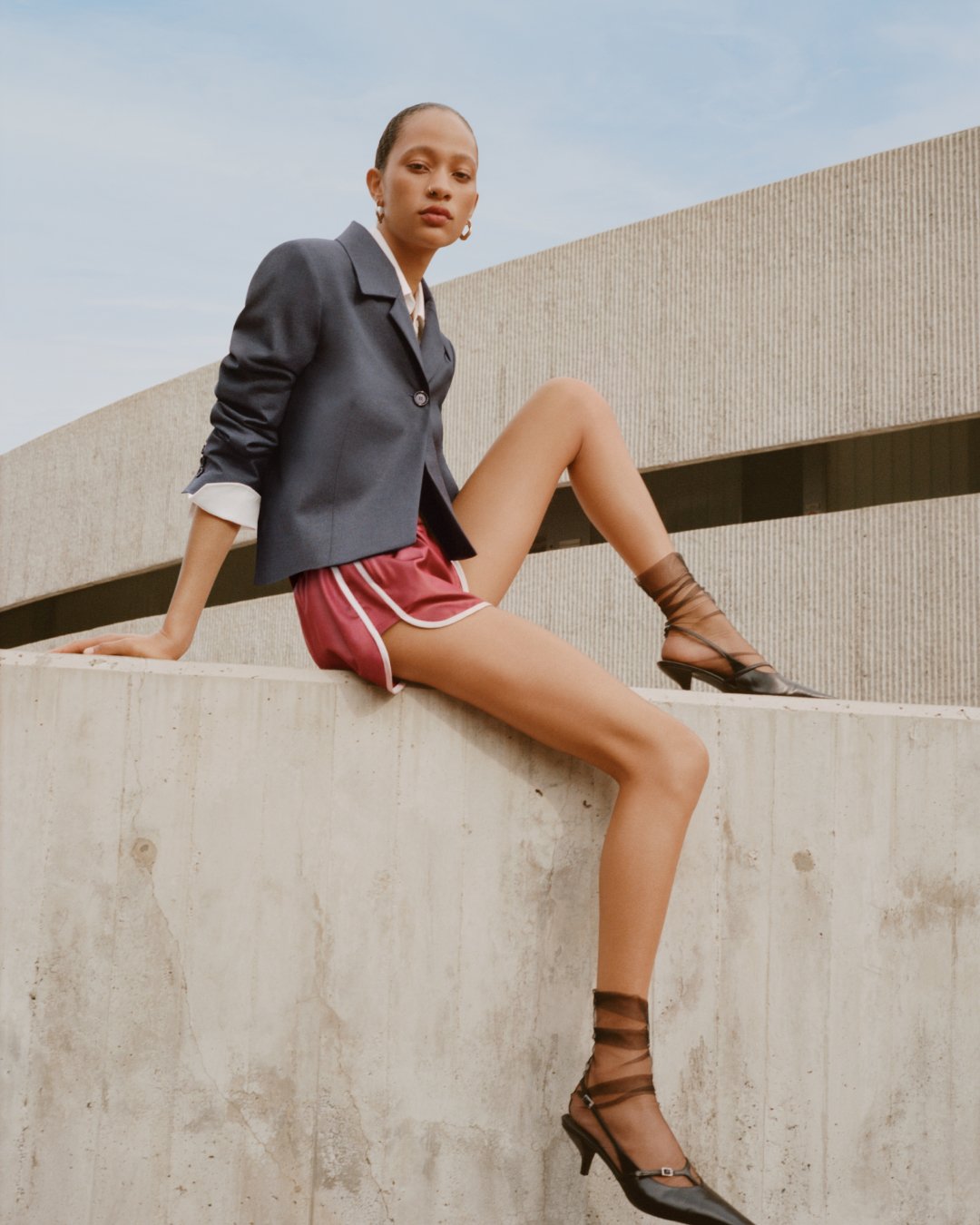 Mango spot - Person sitting on concrete ledge wearing a navy blazer, maroon shorts, and black heels with sheer socks.