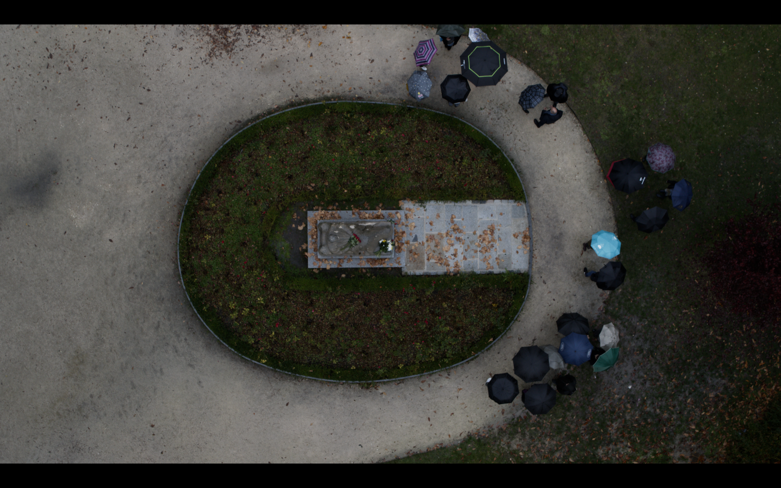 Aerial view of people with umbrellas gathered around a grave in an oval garden.