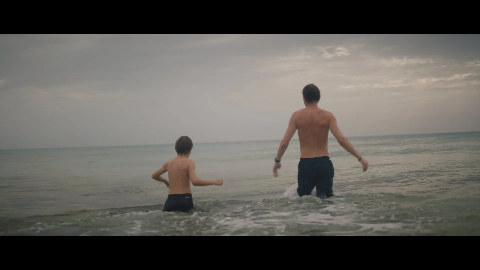Two people wading into the ocean, one adult and one child, both wearing swim shorts, under a cloudy sky.