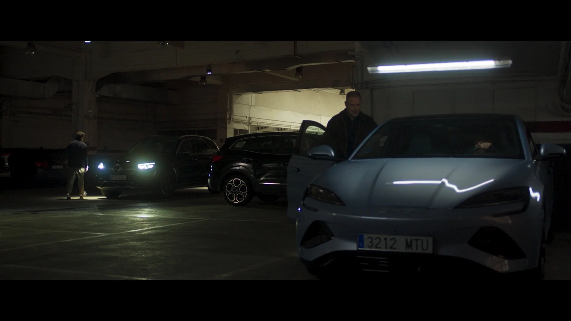 Dimly lit parking garage with parked cars and two people; one standing by a car with headlights on, another entering a black car.