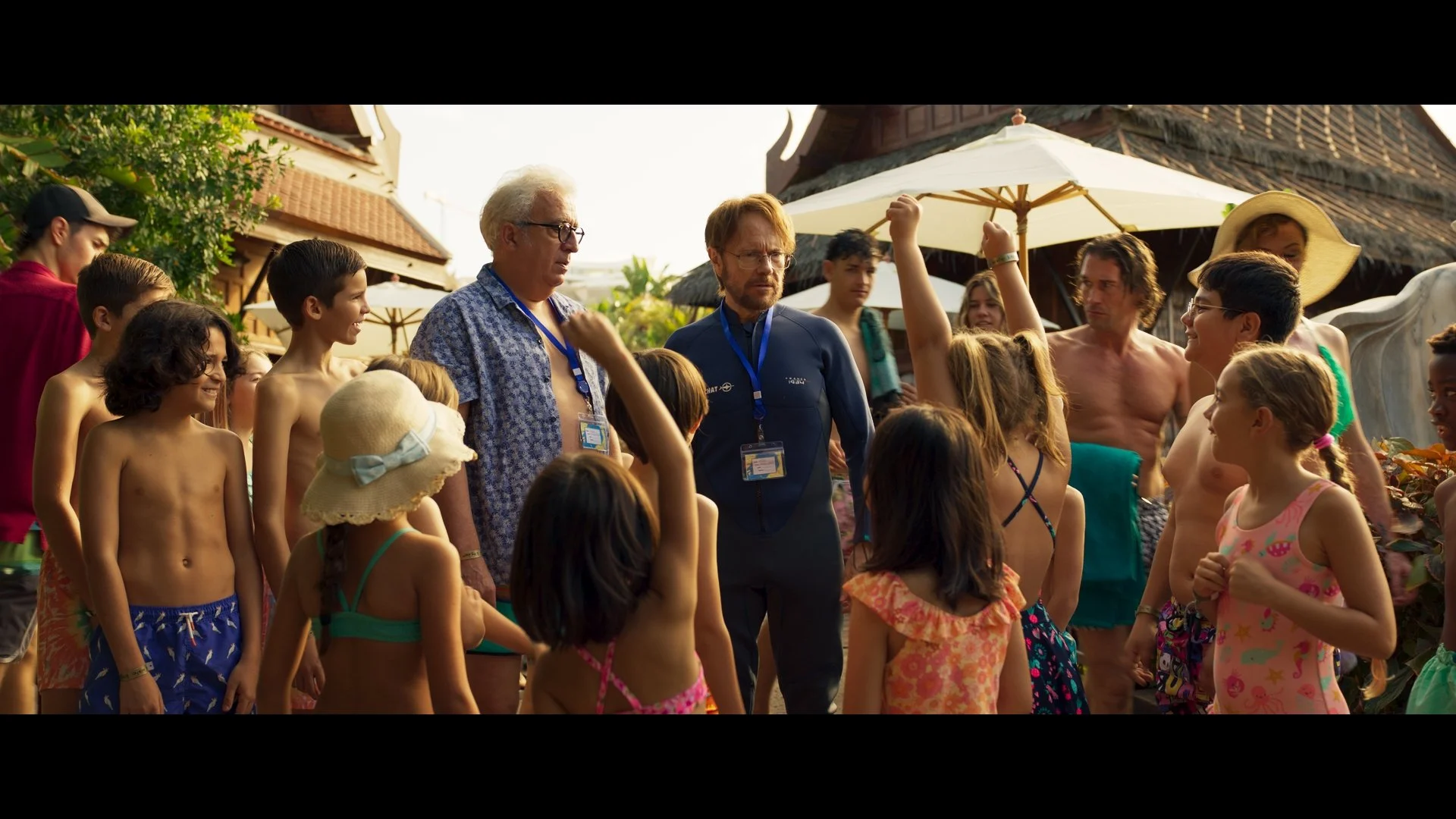 Vacaciones de Verano movie Group of children and adults gathered outdoors, wearing summer clothing, with some children raising hands, under a bright sky.
