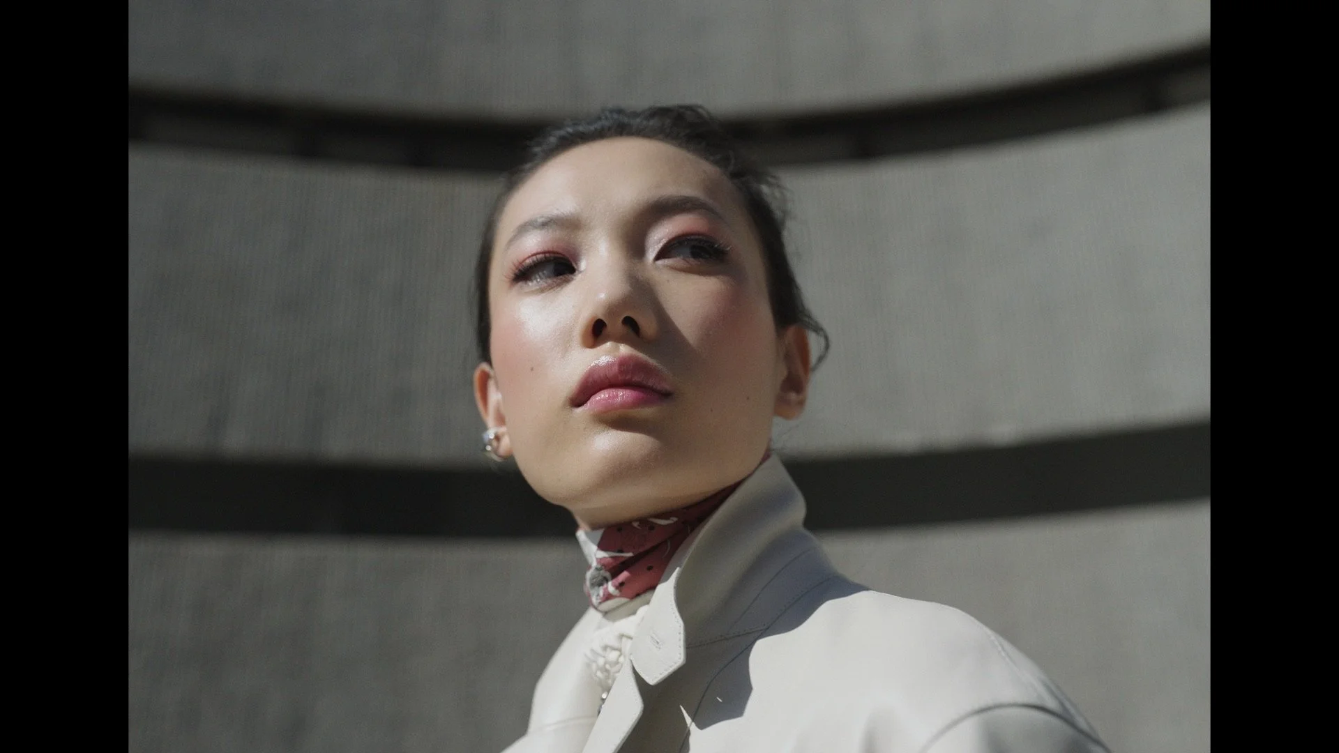 Hermes spot Fashionable person wearing a high-collared jacket and scarf, standing in front of a textured building.