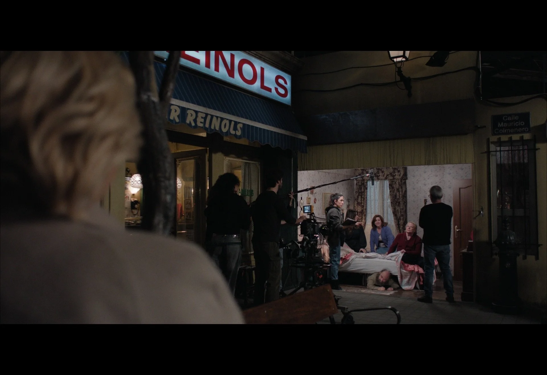 Film crew shooting a scene inside a street-side set, with actors and camera equipment visible. A person in a coat observes from the foreground.