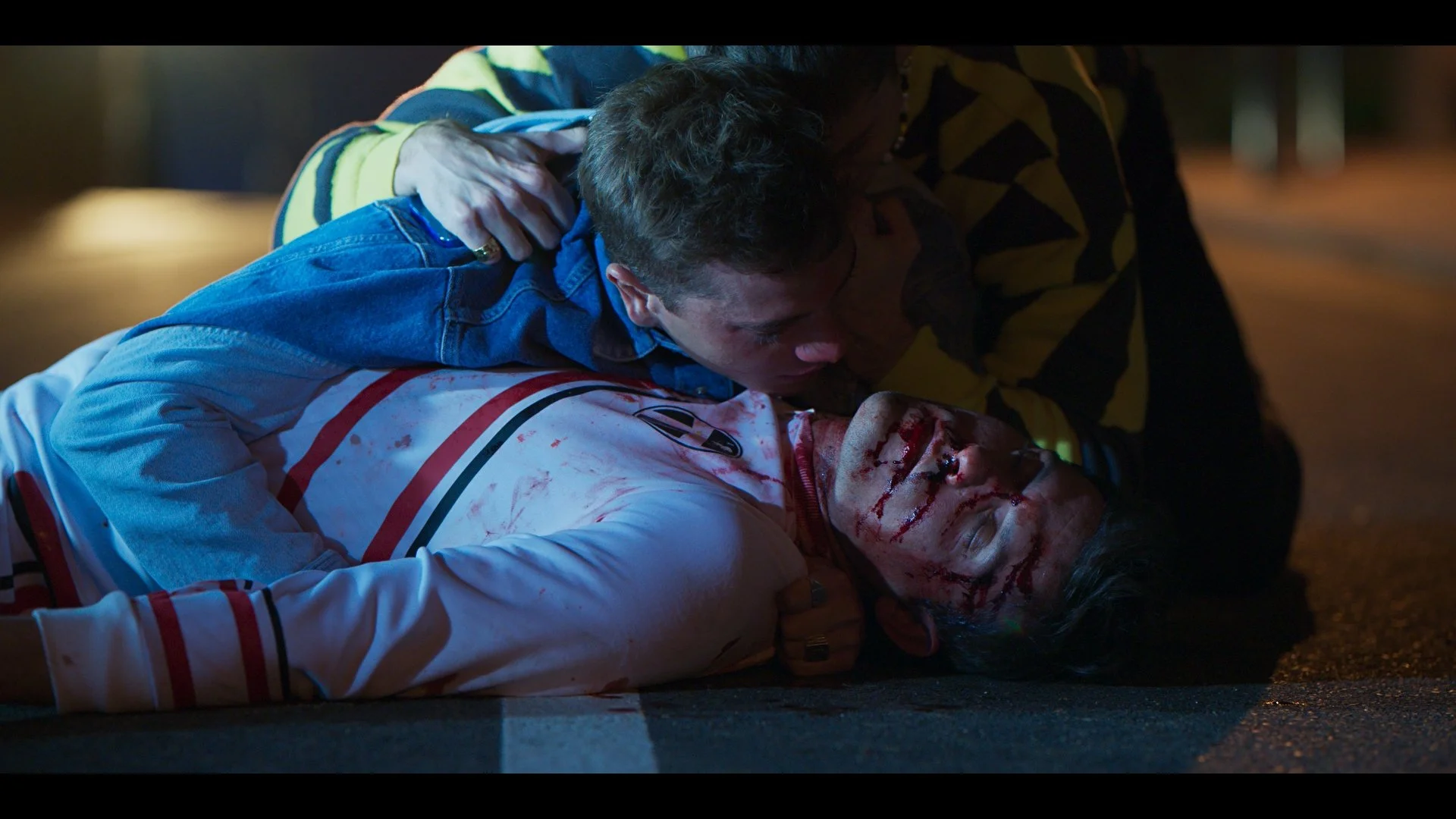 Two men on a dimly lit street, one lying on the ground with a bloodied face, wearing a white shirt with red stripes, and the other leaning over him, appearing concerned.