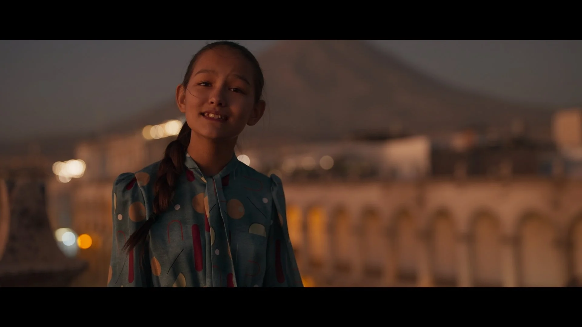 A girl with braided hair wearing a colorful, patterned dress stands outdoors at dusk with blurred city lights in the background.