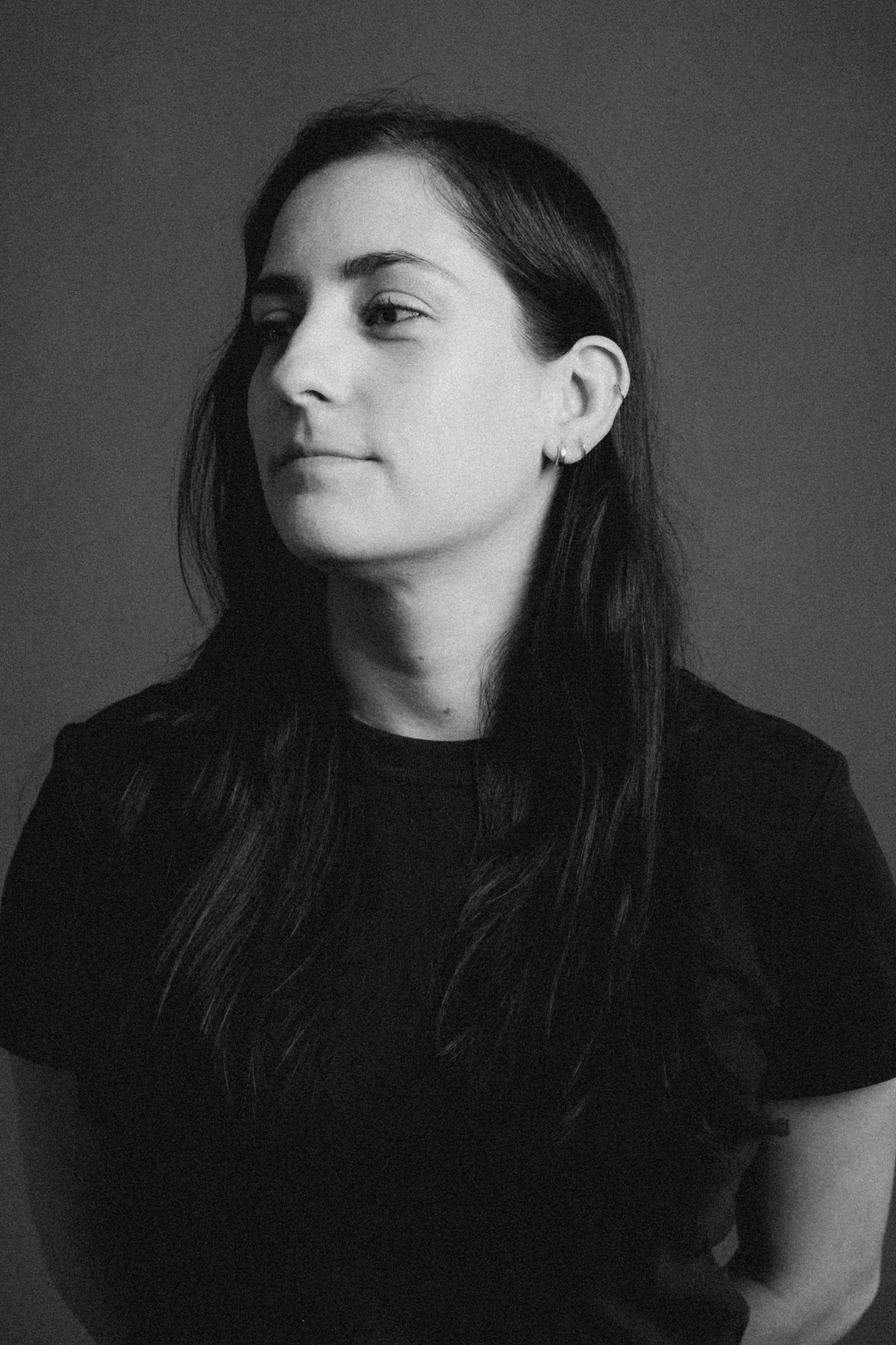 Retrato en blanco y negro de una mujer con cabello largo y oscuro, usando orejeras pequeñas y una camisa negra, mirando a la izquierda con expresión neutra.