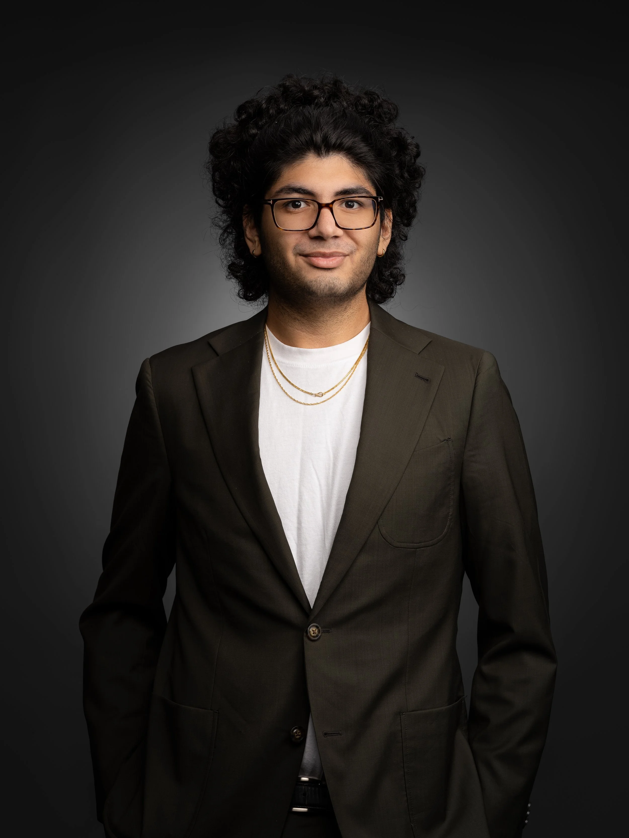 Man with glasses wearing a dark suit jacket and white shirt in a studio portrait.