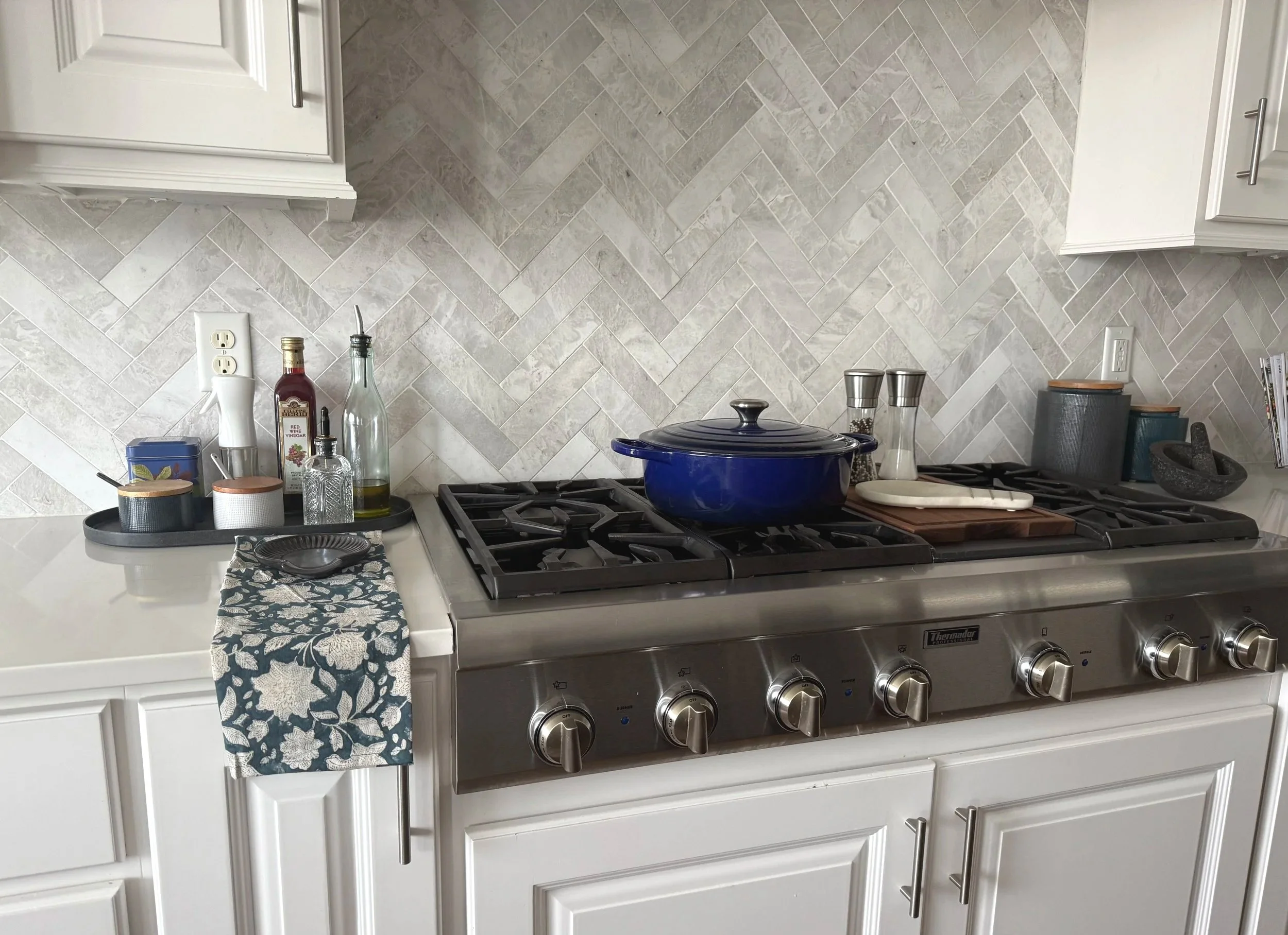 Kitchen stove with marble herriingbone pattern backsplash.  White snowbound cabinets above and a patterned cloth hanging on the side.