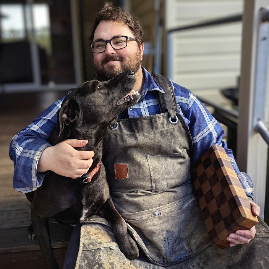 A man with glasses and a beard, wearing a blue plaid shirt and an apron, sitting outside with a black dog on his lap. The dog is looking up and licking the man's face. The man is holding a checkered wooden box in his right hand and smiling.