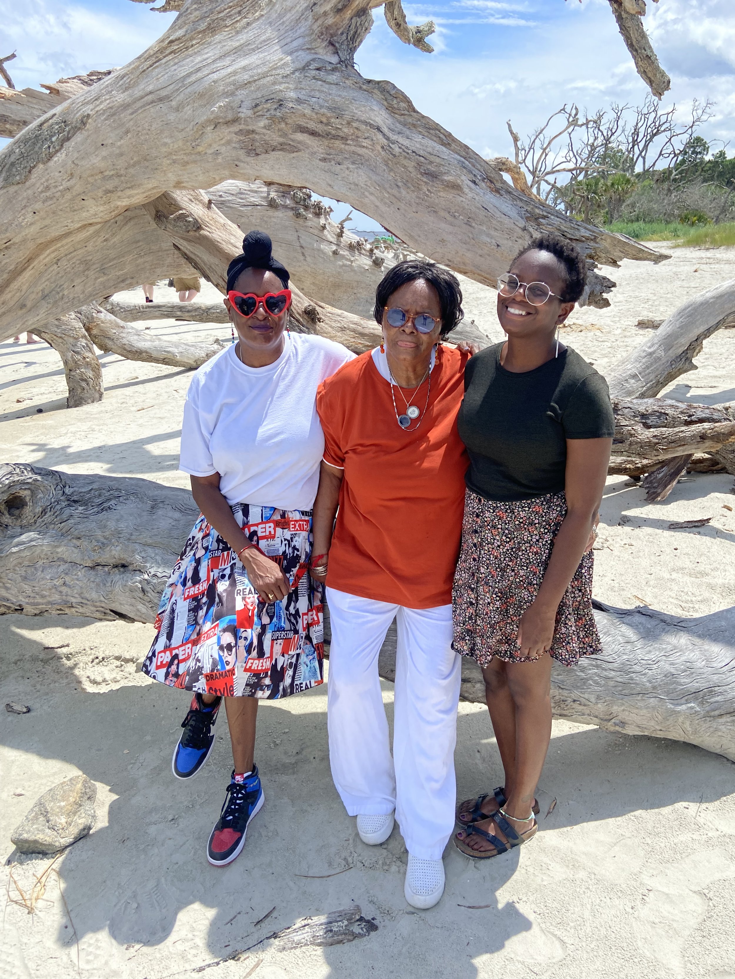 Three women standing on a sandy beach, smiling in front of a large, weathered driftwood. The sky is clear and blue, and there are some trees in the background. They are wearing casual summer attire, including skirts and sunglasses.