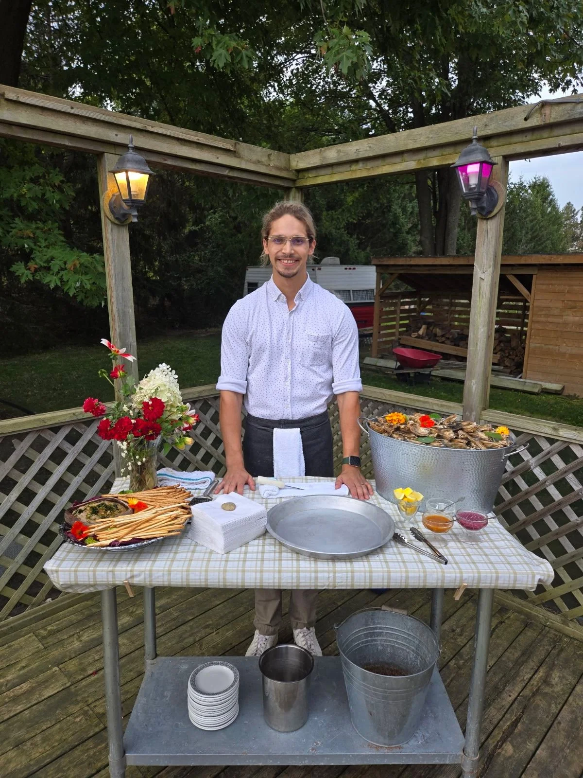 A young man with glasses is standing behind a table outdoors, preparing seafood with lemons, sauces, and a large tub of oysters, at a backyard deck with a wooden fence and trees in the background.