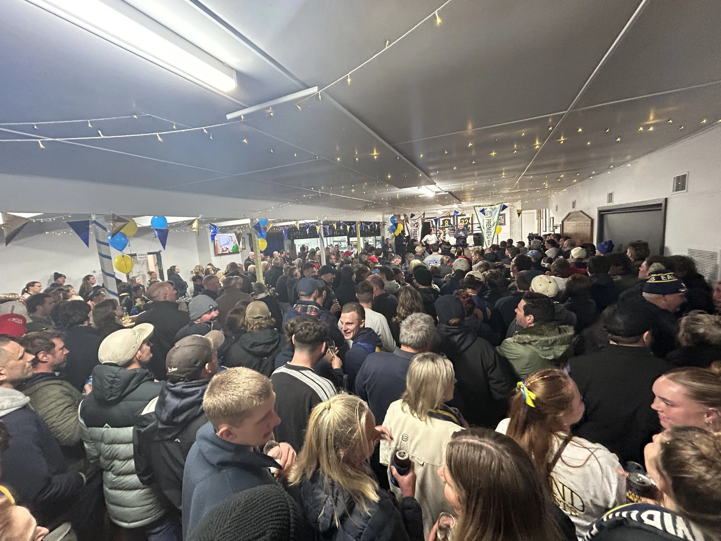 Crowd of people gathered in a large indoor space decorated with blue and yellow balloons and banners, watching a stage at the front.