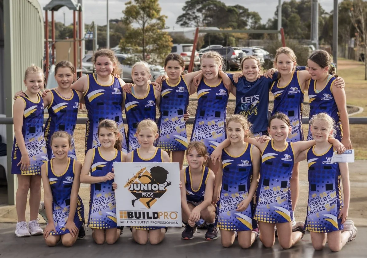Group of young girls in blue and yellow sports uniforms posing outdoors on a sports field, holding a sign that says 'Junior Pros' and 'Build Pro.'