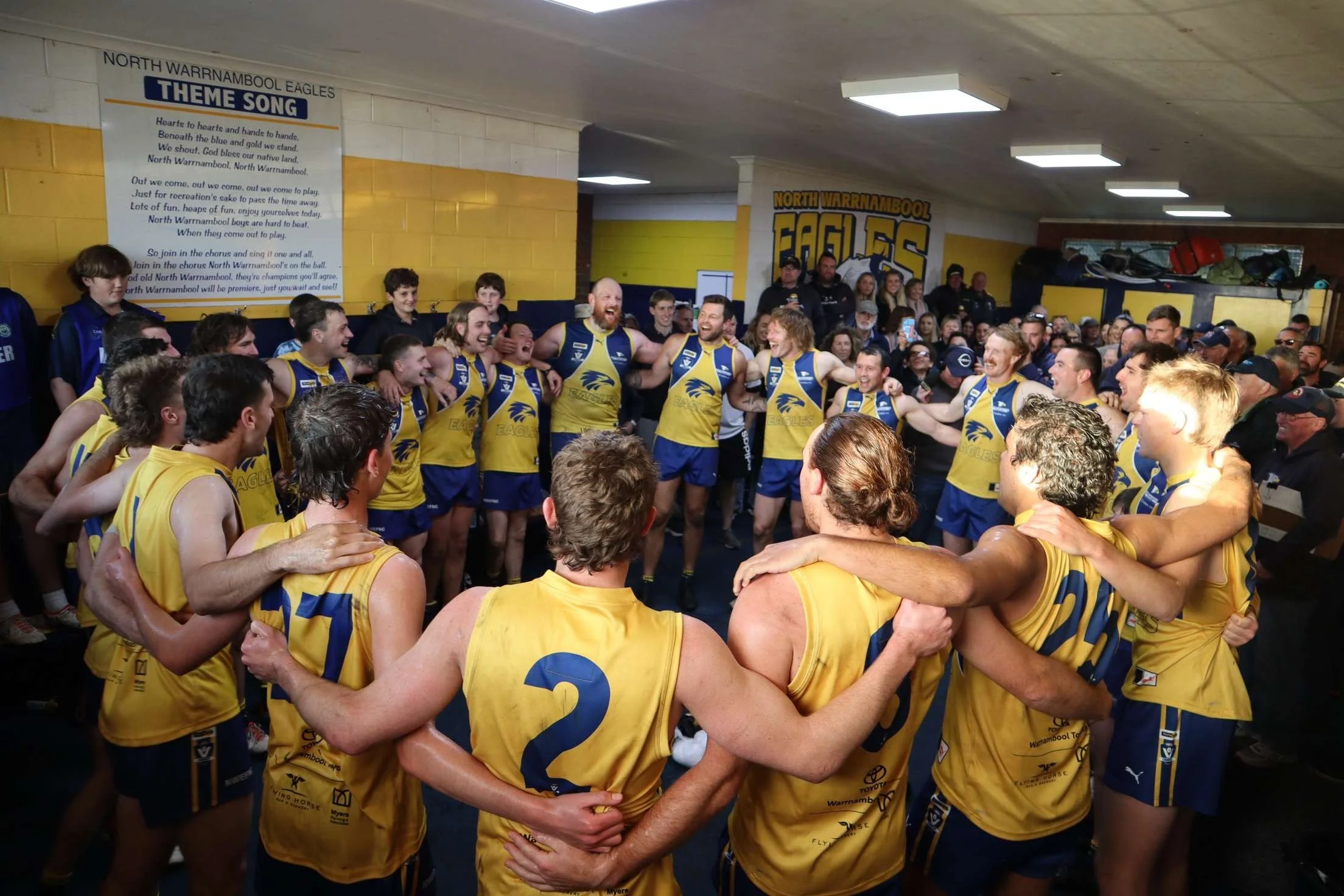Australian Rules football team in a dressing room, celebrating in a circle after a game, wearing yellow and blue uniforms with the team logo.