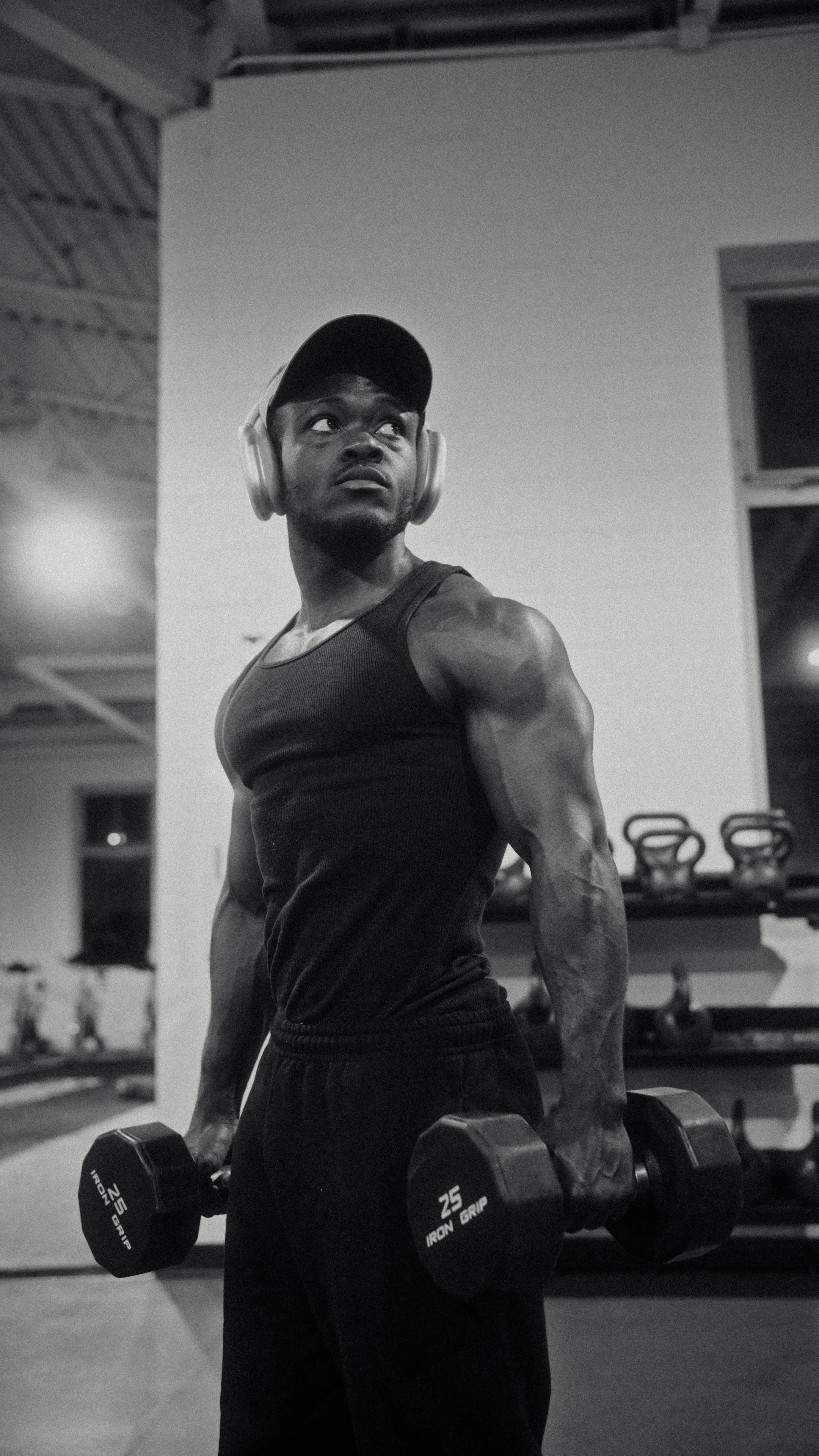 A man in a gym holding dumbbells in both hands, wearing a cap, headphones, and a sleeveless shirt.