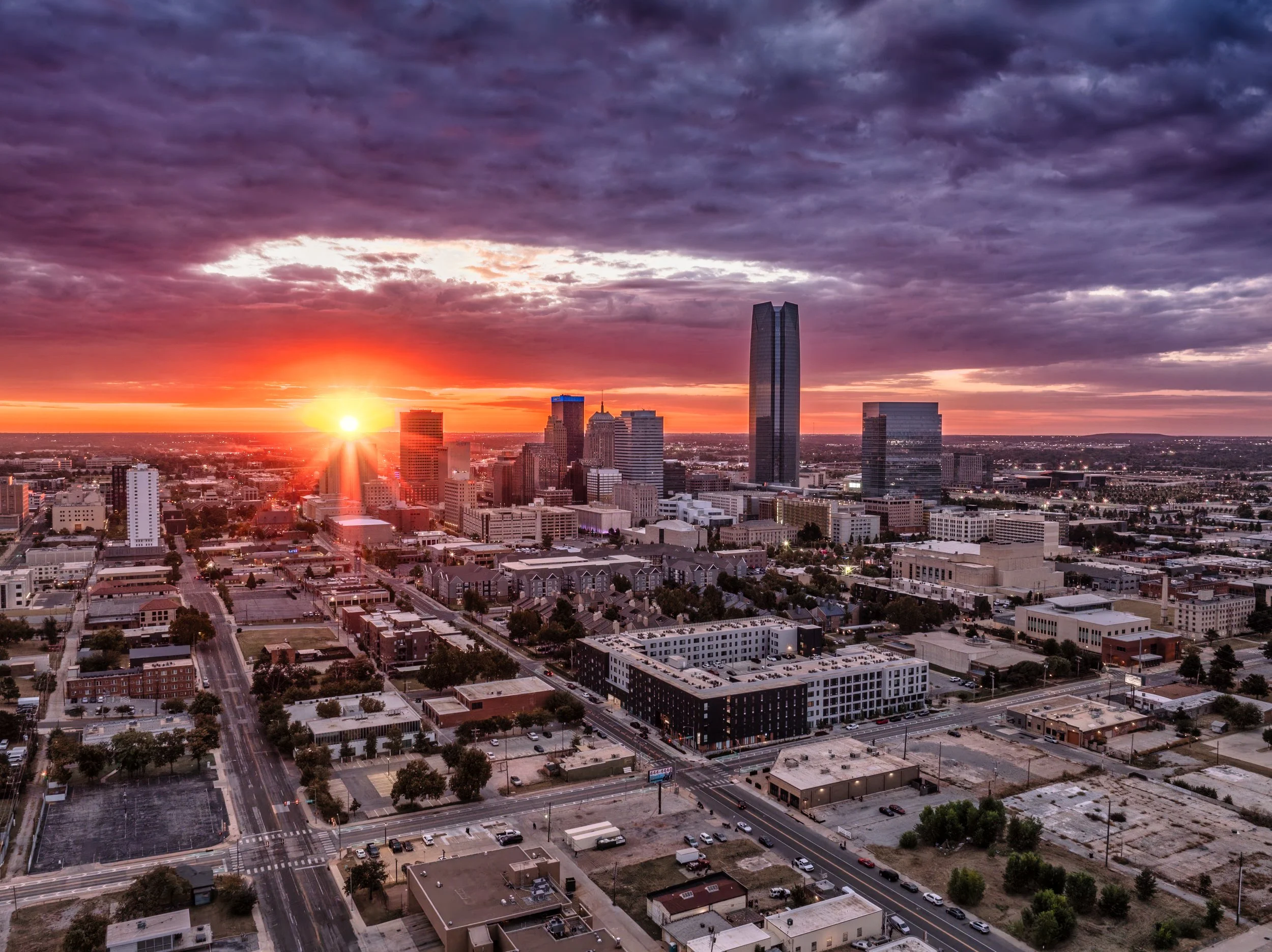A city skyline during sunset with tall skyscrapers, with one prominent dark glass building, under a dramatic sky with clouds and the setting sun near the horizon.