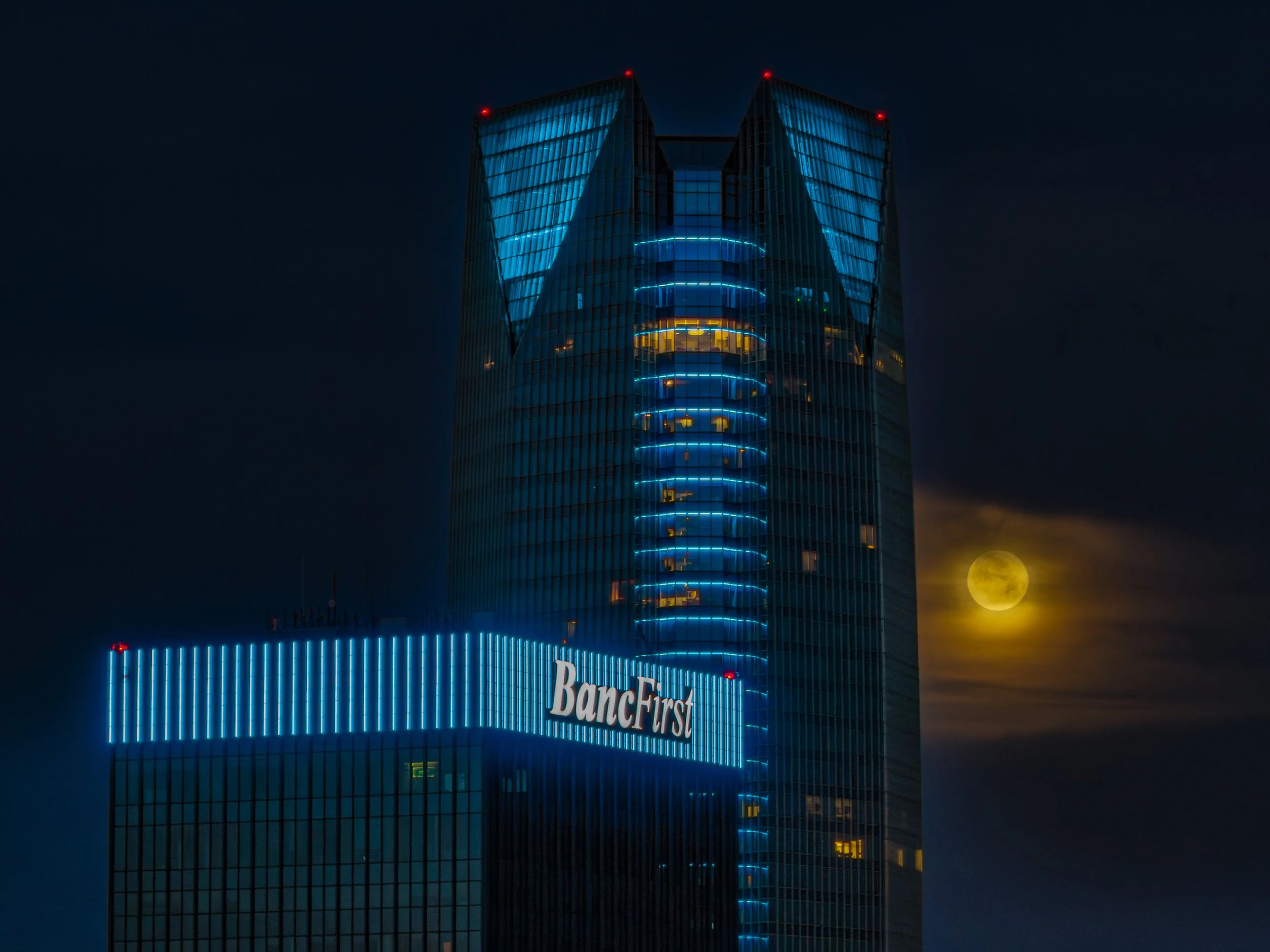 Nighttime photo of a tall glass skyscraper with blue LED lights and a bright yellow full moon in the background. The building has a sign that reads 'BancFirst'.