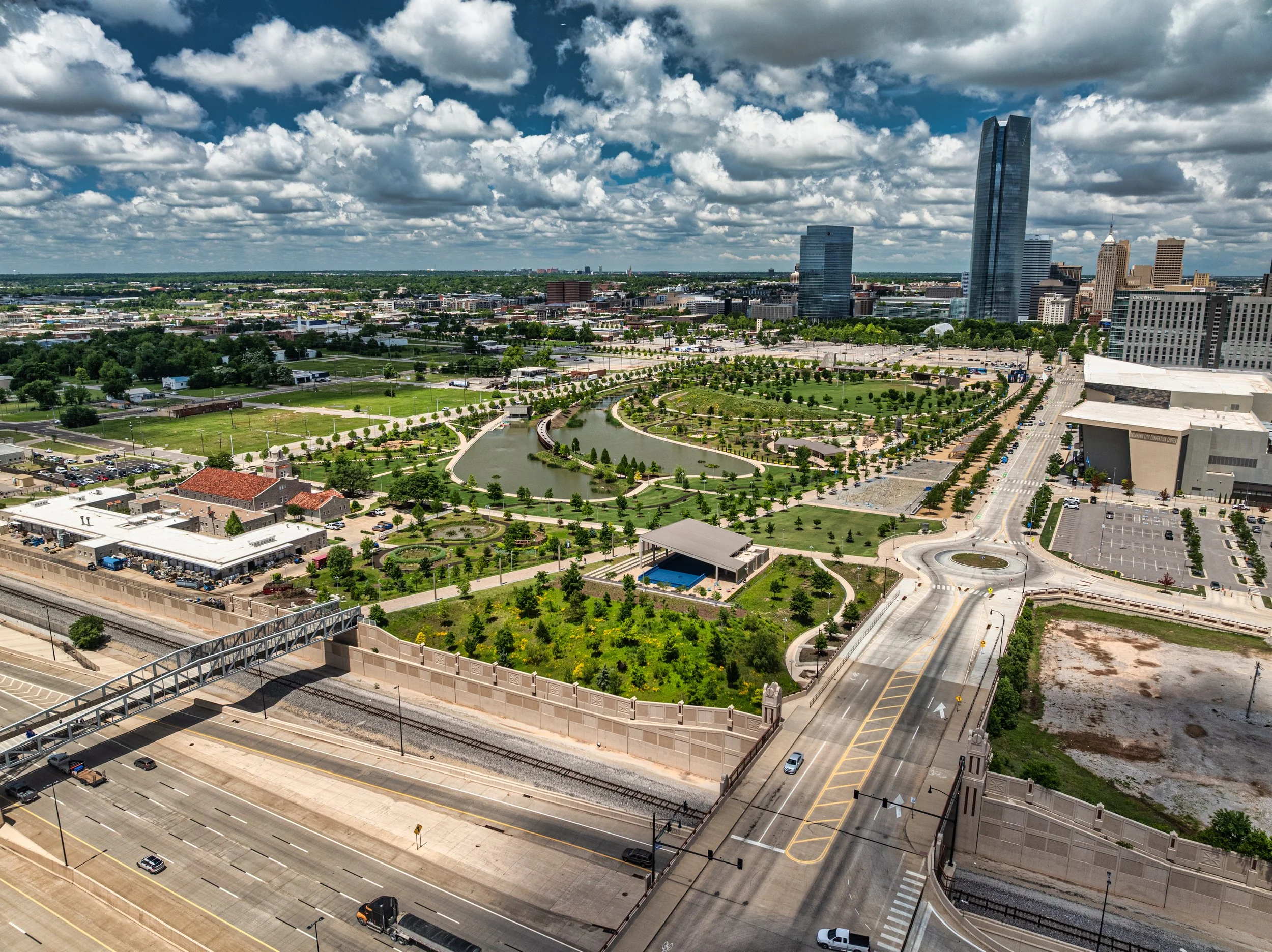 Aerial view of a city park with walking paths, green trees, and a small pond, surrounded by roads, buildings, and skyscrapers under a partly cloudy sky.