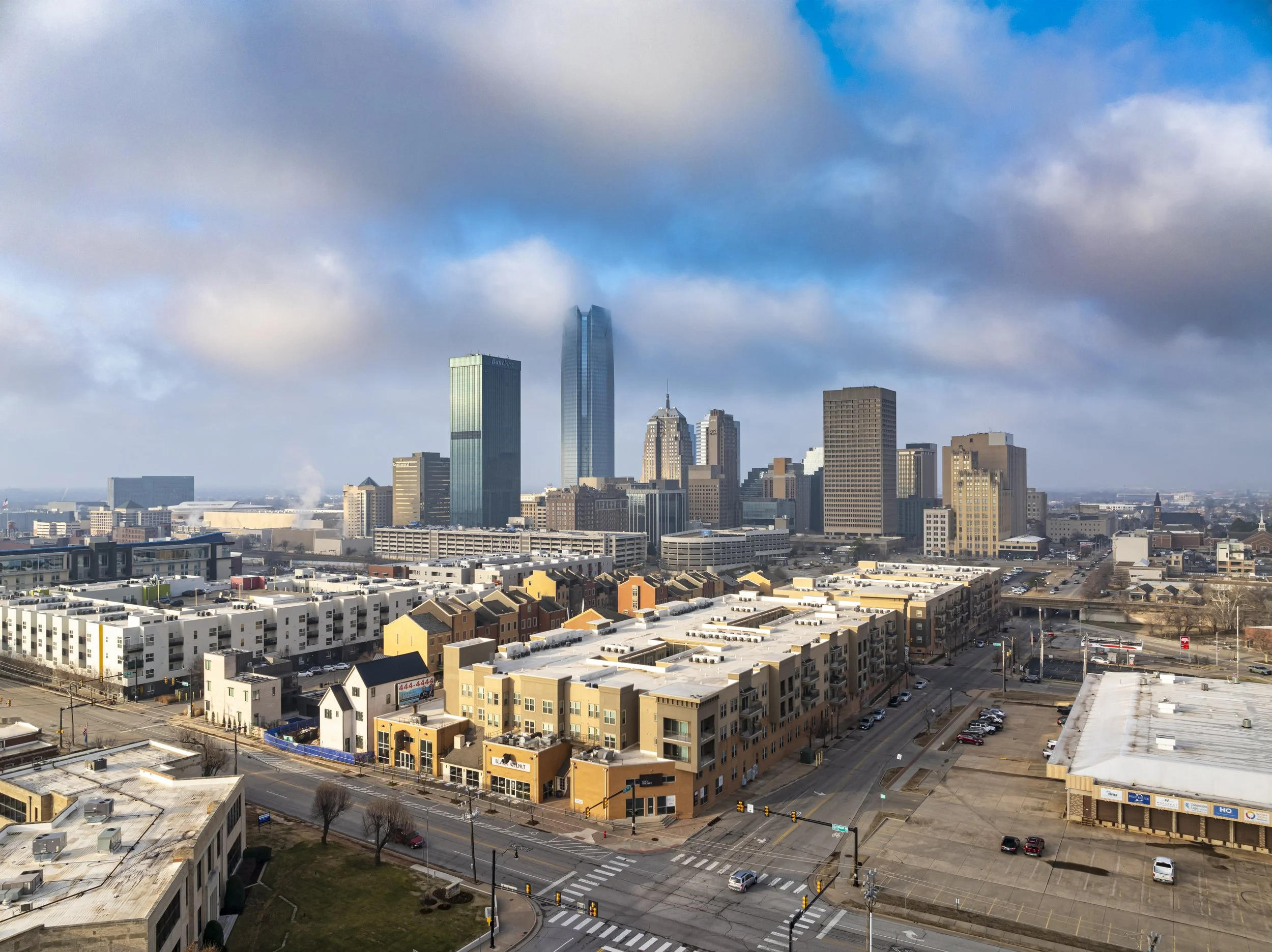 City skyline with tall skyscrapers behind a mix of residential and commercial buildings, under a partly cloudy sky.