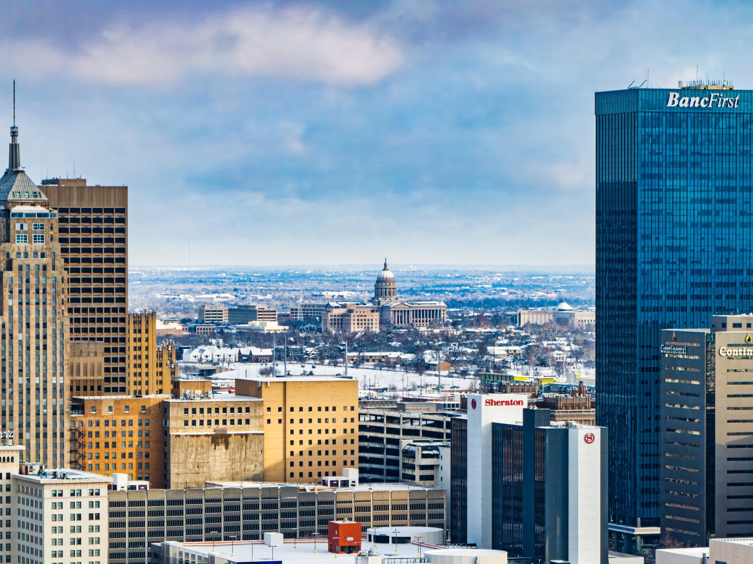 City skyline with tall buildings, including a blue glass skyscraper, and a distant view of a domed government building, under a partly cloudy sky with snow-covered ground.