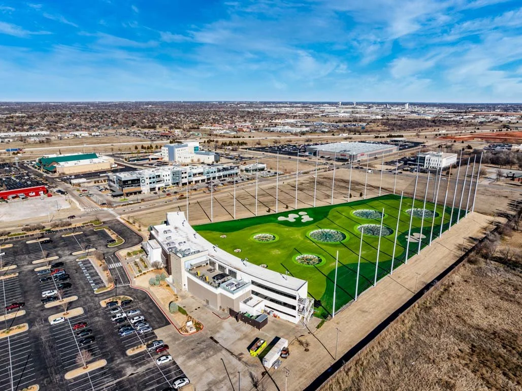 An aerial view of a golf practice area with multiple miniature golf holes, a white clubhouse, and a parking lot, with commercial buildings and an open landscape in the background under a partly cloudy sky.