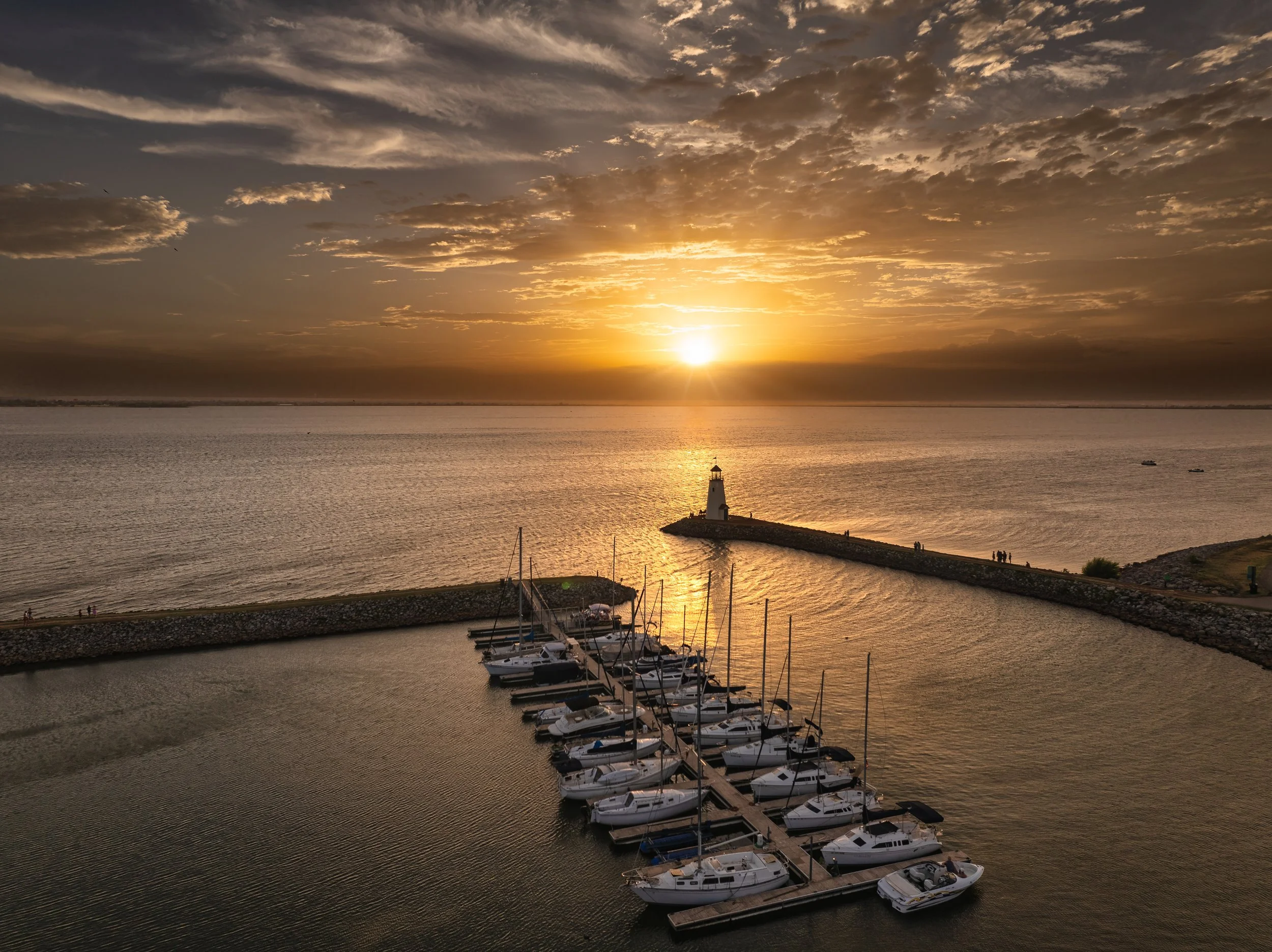 Sunset over a marina with several sailboats docked at the pier. A lighthouse on a breakwater extends into the water, with a small group of people nearby. The sky is partly cloudy with the sun visible near the horizon, casting a golden light.