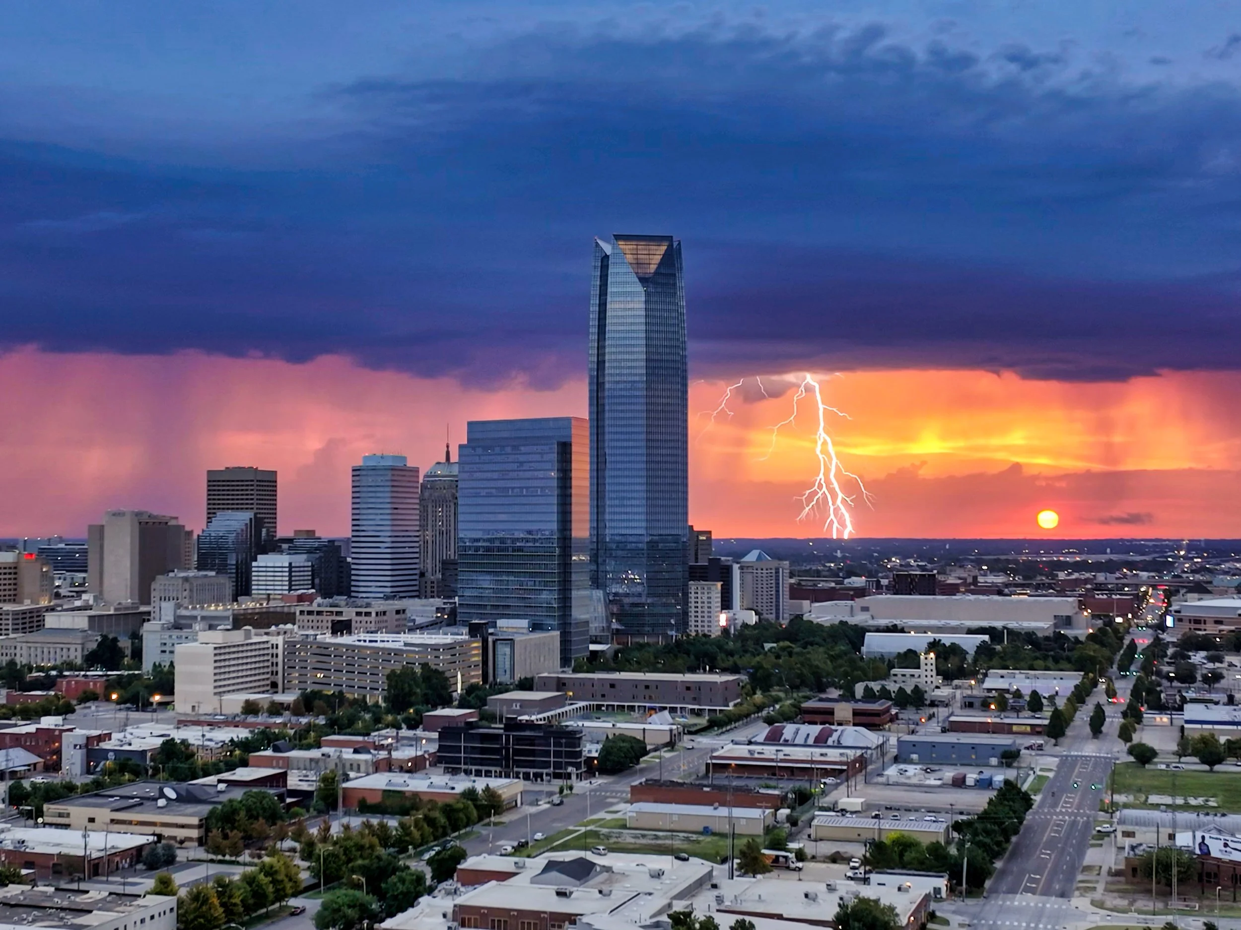 City skyline at sunset with dark storm clouds, lightning in the sky, and the sun near the horizon.