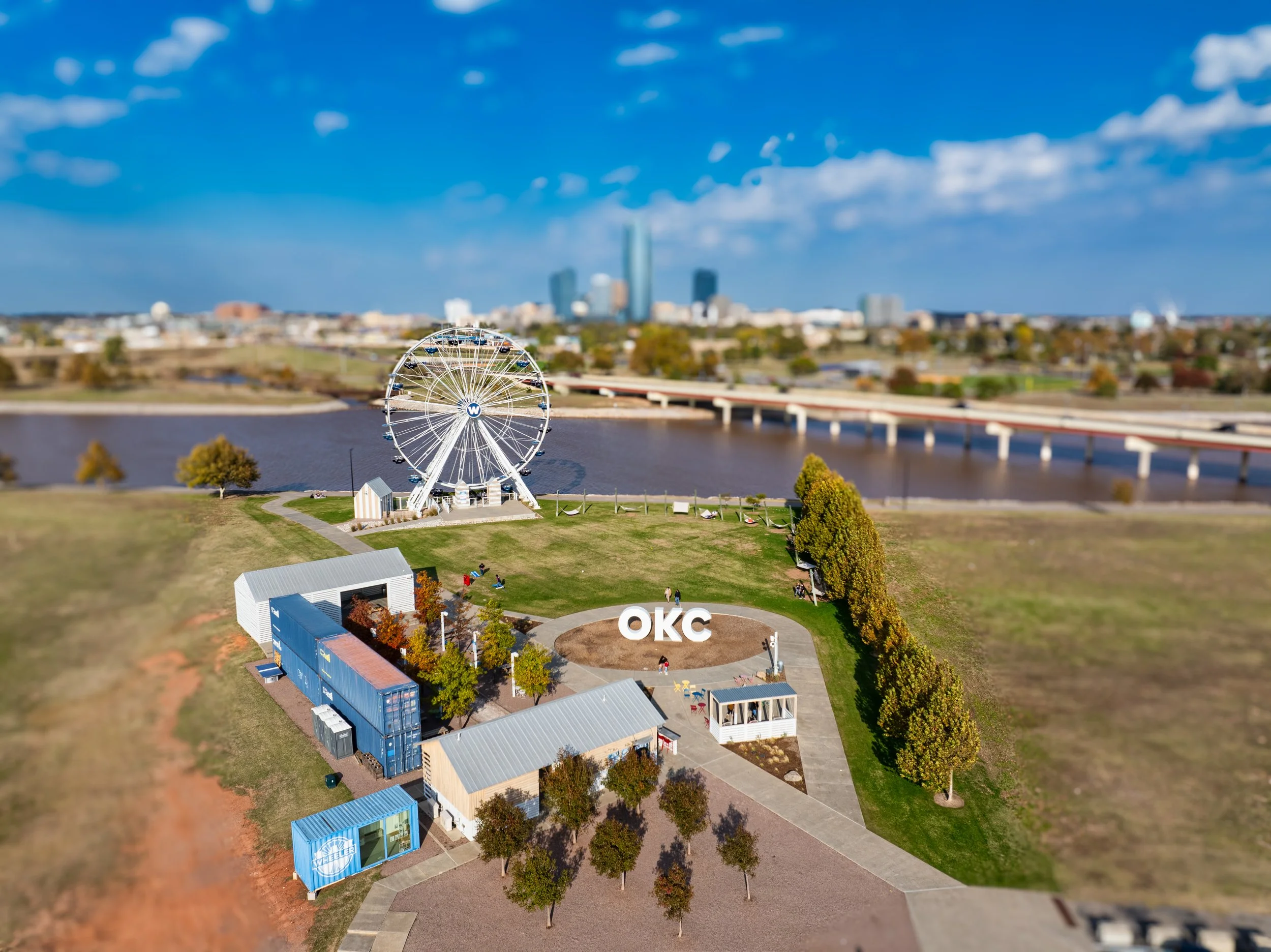 Aerial view of a park in Oklahoma City with an illuminated Ferris wheel, large OKC sign, and shipping containers near trees, with a river and city skyline in the background.