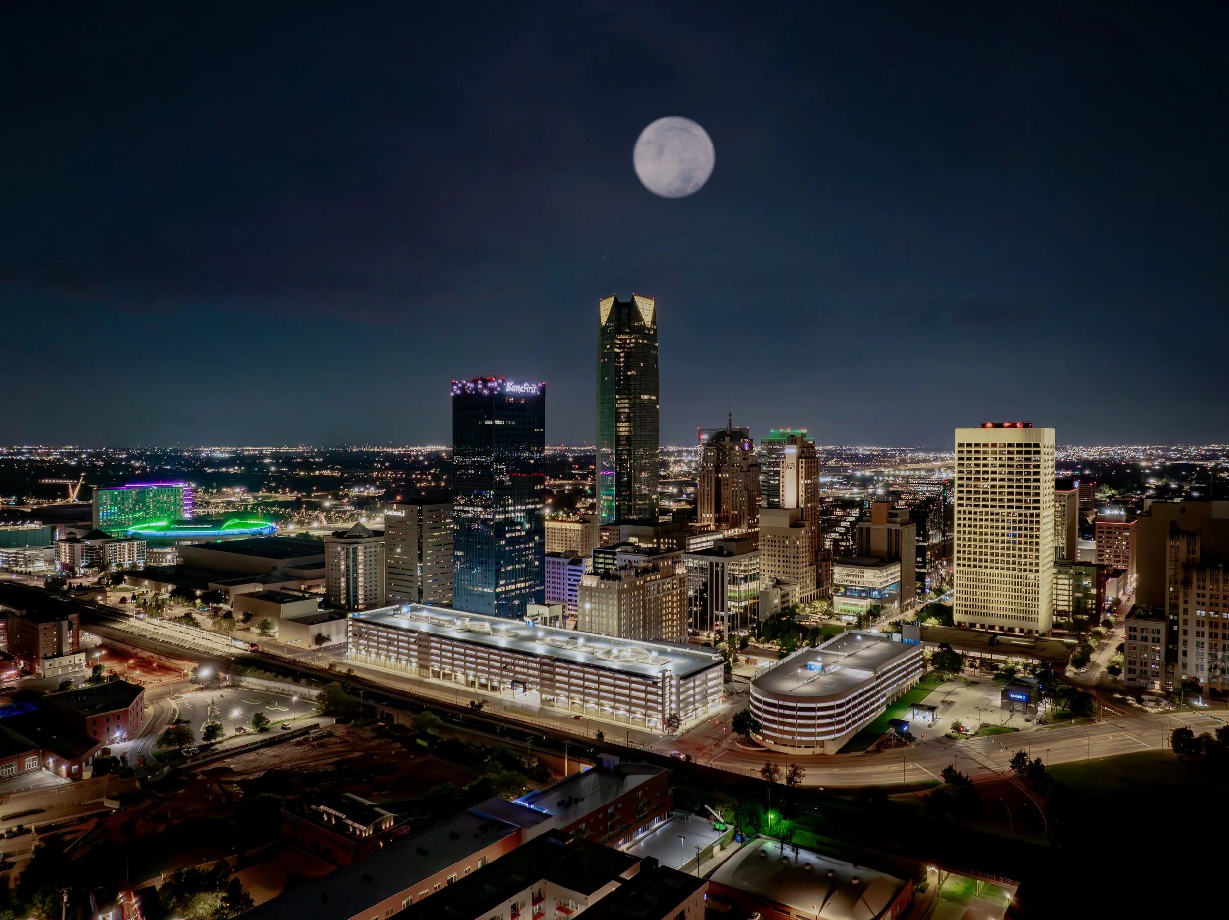 Nighttime city skyline with tall buildings, illuminated streets, and a full moon.