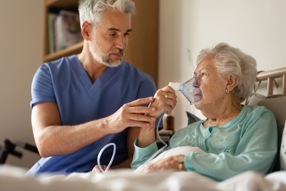 Male nurse helping an elderly woman use an oxygen mask, delivering respiratory support through home nursing care in Australia.