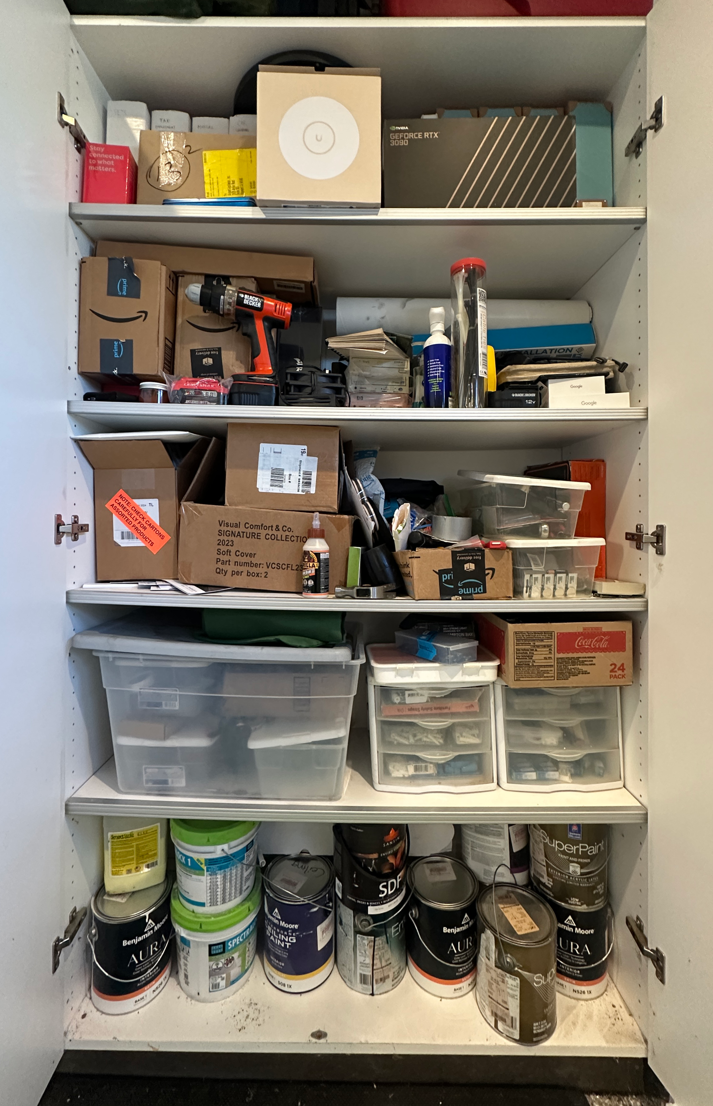 Before of a white garage cupboard that is semi disorganized and includes white tubs and many gallons of paint.