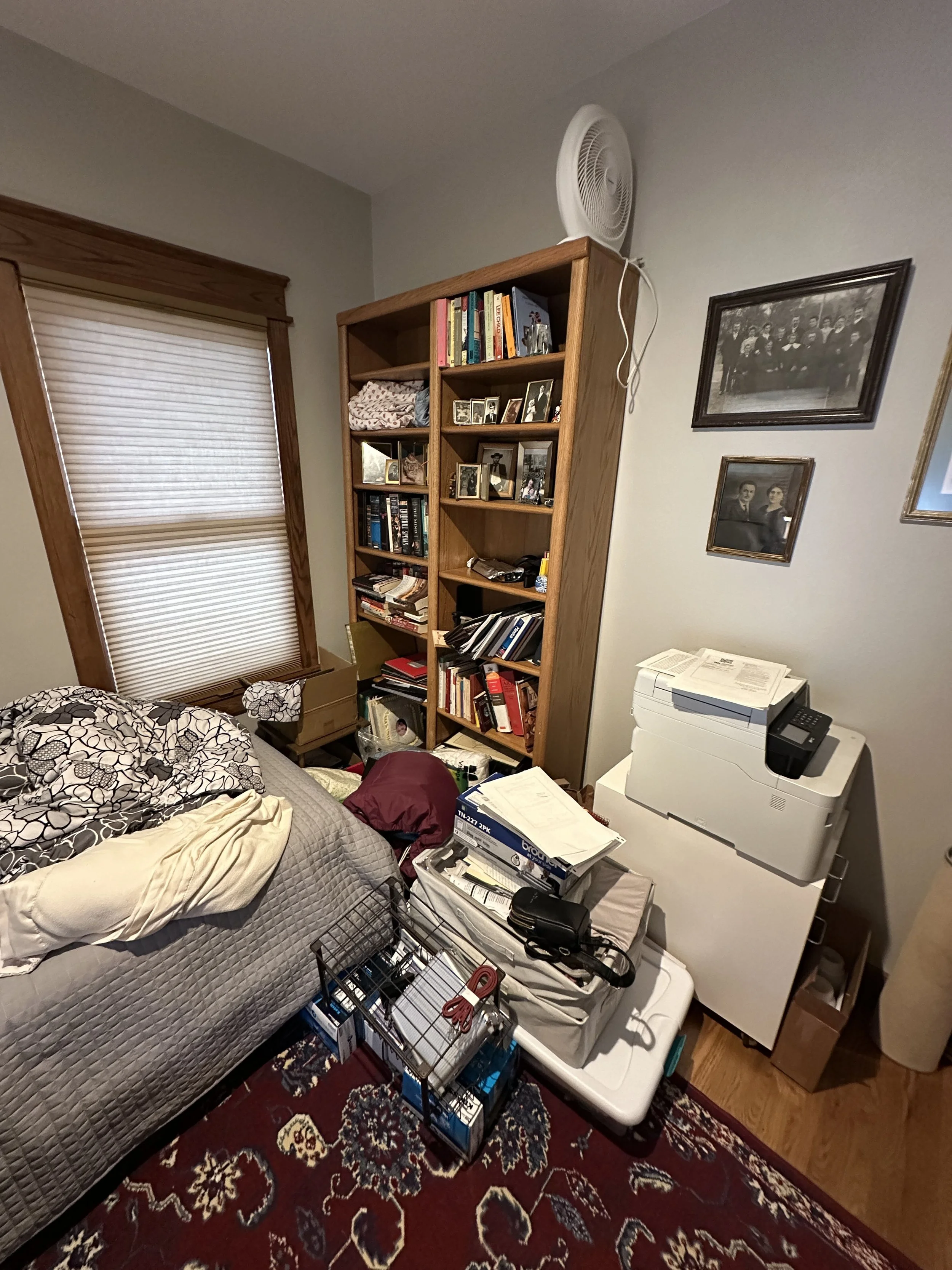 Before image of a spare bedroom with piles of nostalgic items and paperwork on the floor, with a disorganized bookcase in the background.