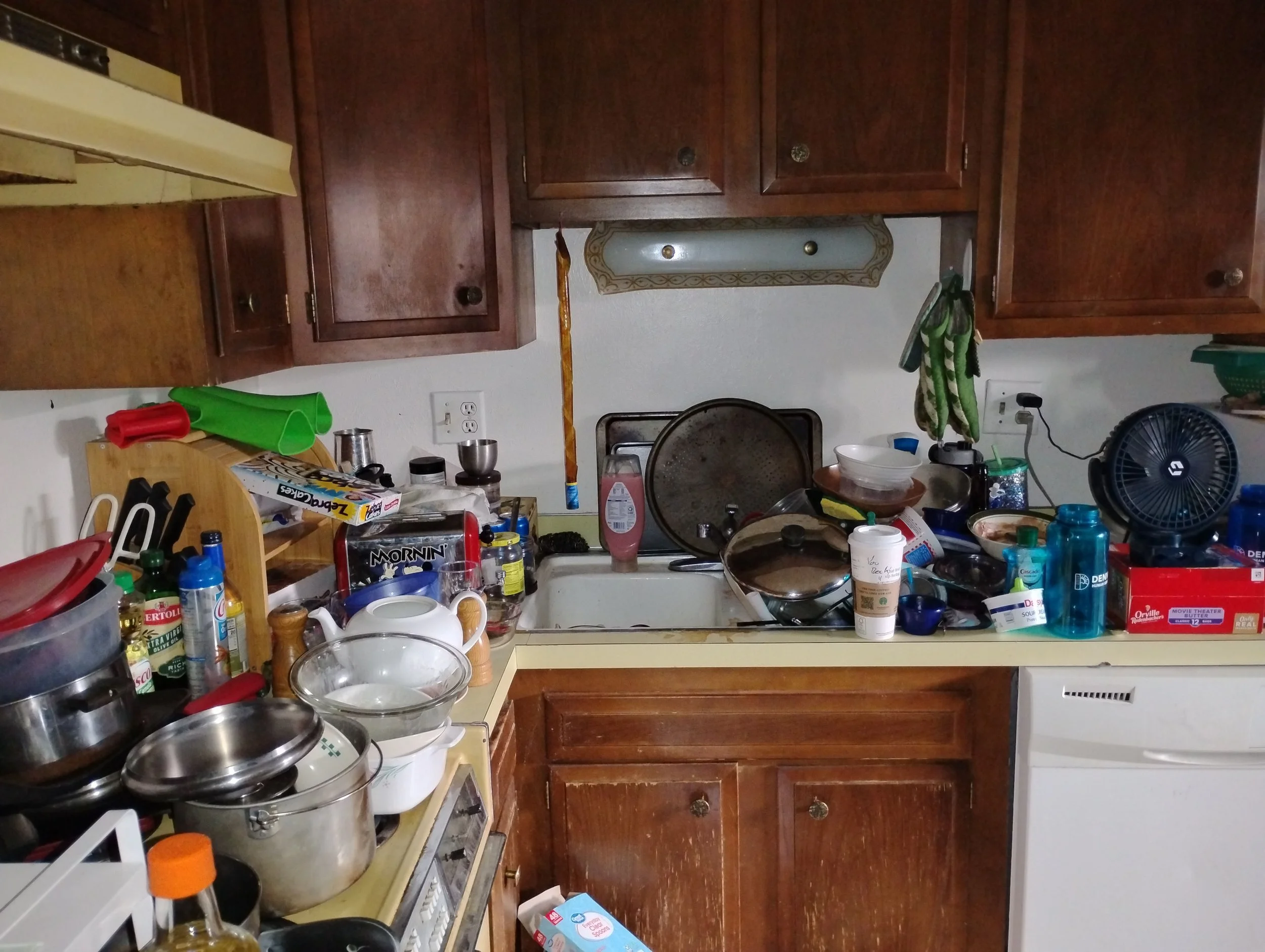Before image of a cluttered kitchen with piled dishes and trash on all horizontal surfaces.