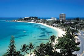 A scenic view of a coastal beach with turquoise waters, white sand, palm trees, and buildings in the background under a clear blue sky.