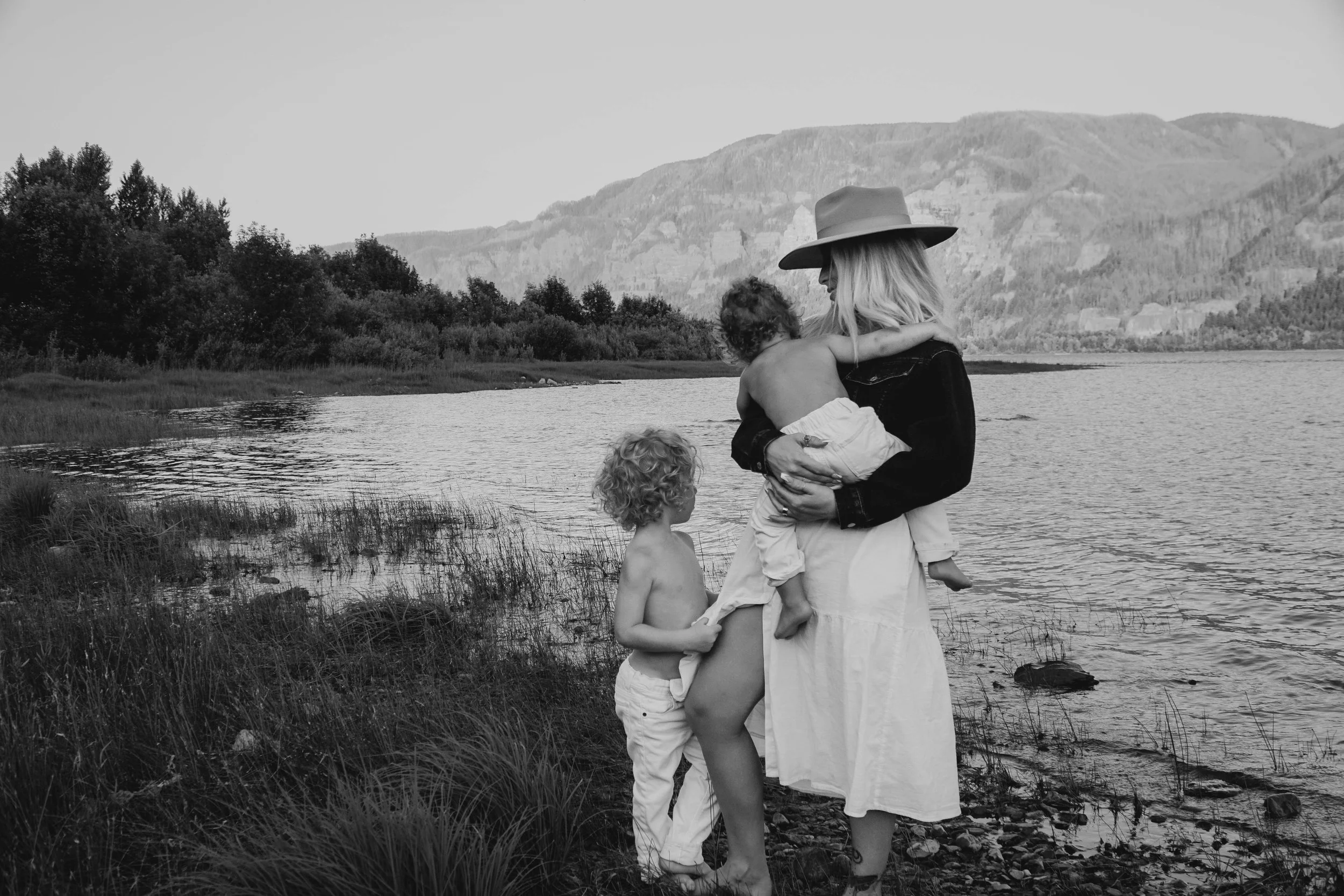 A woman holding a young child while a boy stands nearby by a lakeshore with mountains in the background.