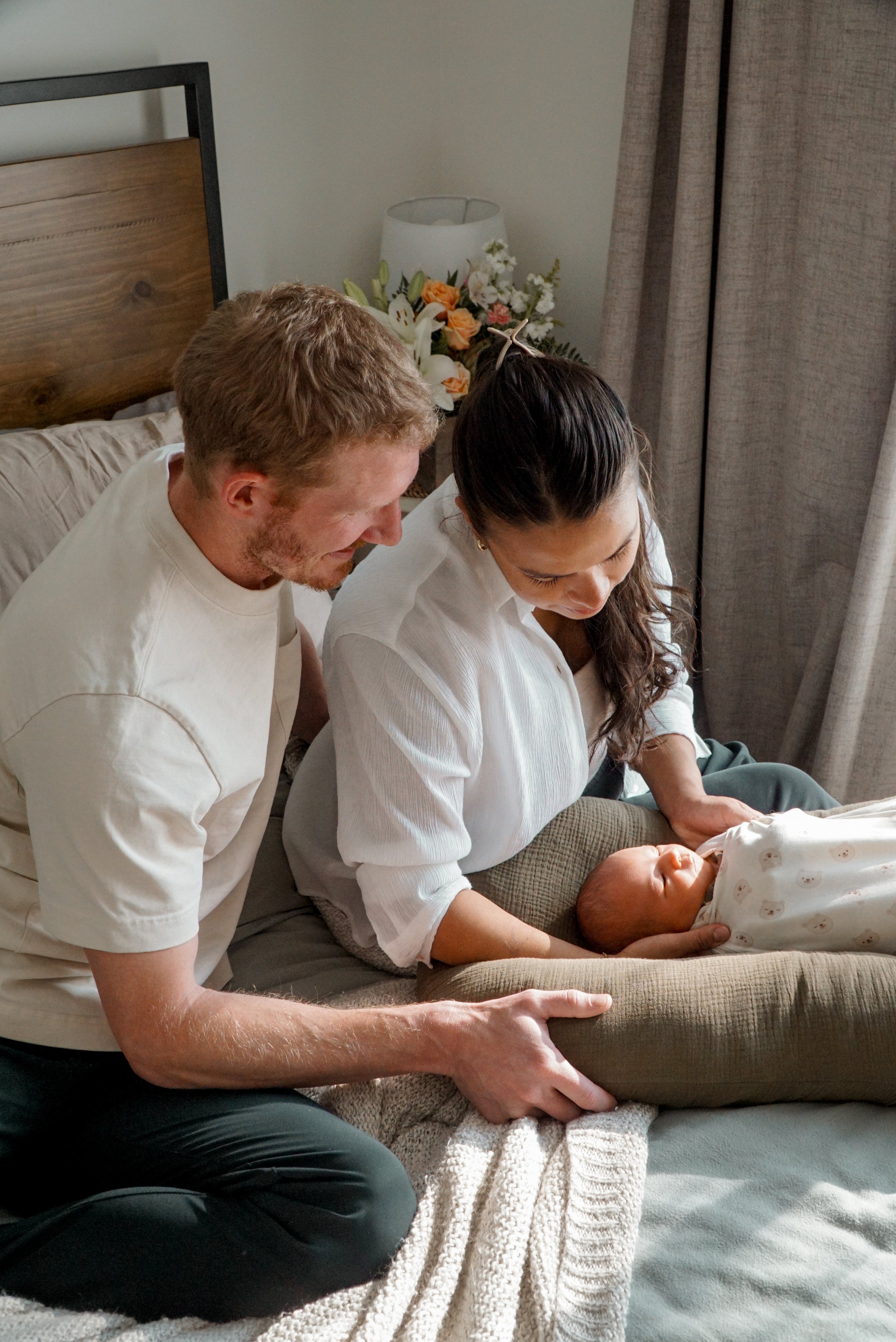 A couple with light skin and dark hair, the woman with long hair in a bun, looking at a sleeping newborn in a crib. The man has short hair and beard. The scene is in a bedroom with flowers and curtains in the background.