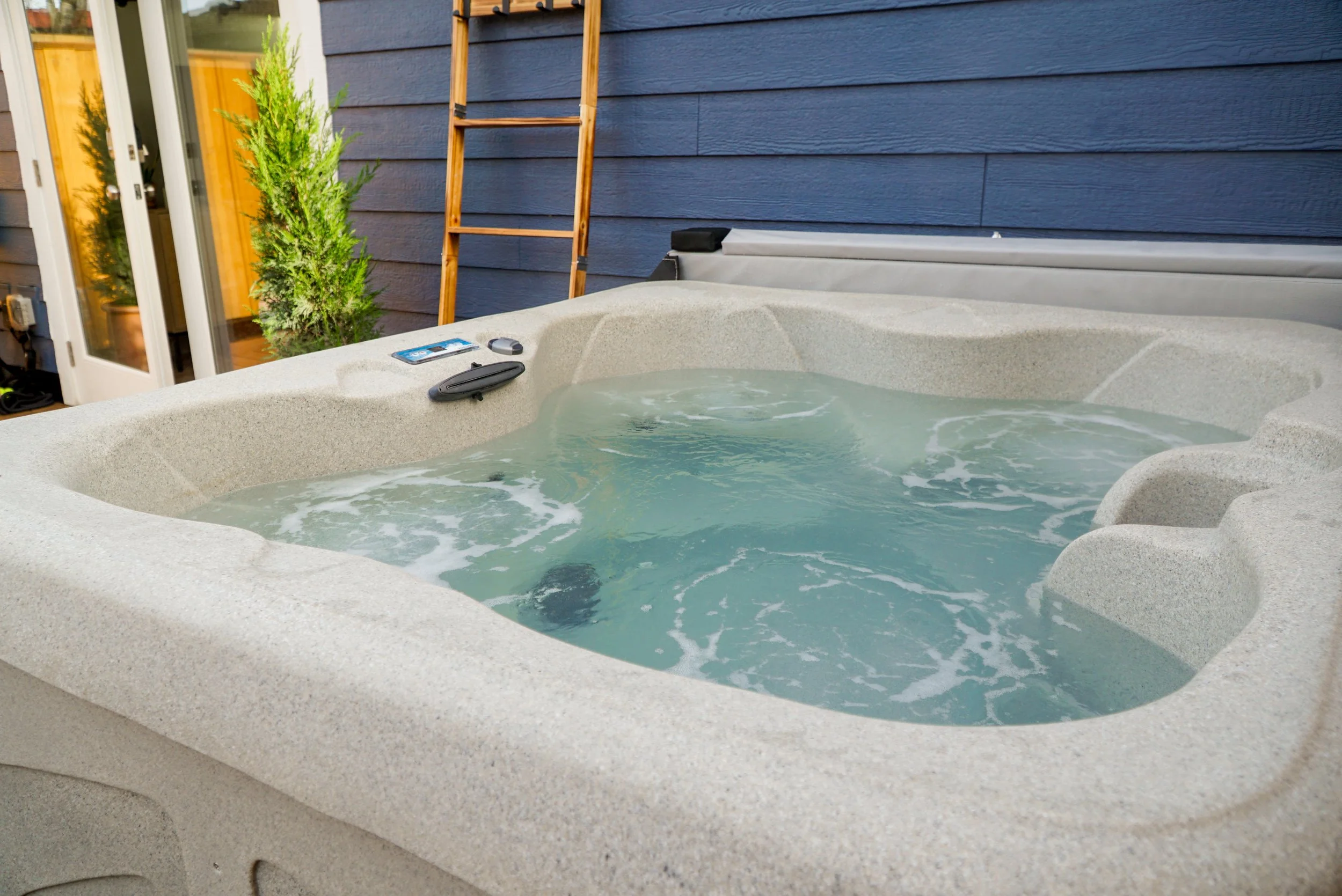 A hot tub filled with bubbling water outdoors next to a blue house with a glass door, a small green plant, a wooden ladder, a remote control, and a phone placed on the hot tub's edge.