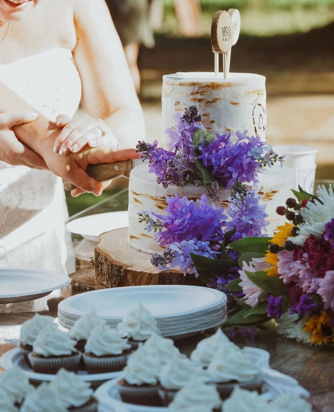A wedding cake with purple flowers and a wooden 'Mrs' cake topper, being cut by the bride, surrounded by plates, cupcakes, and colorful flowers.