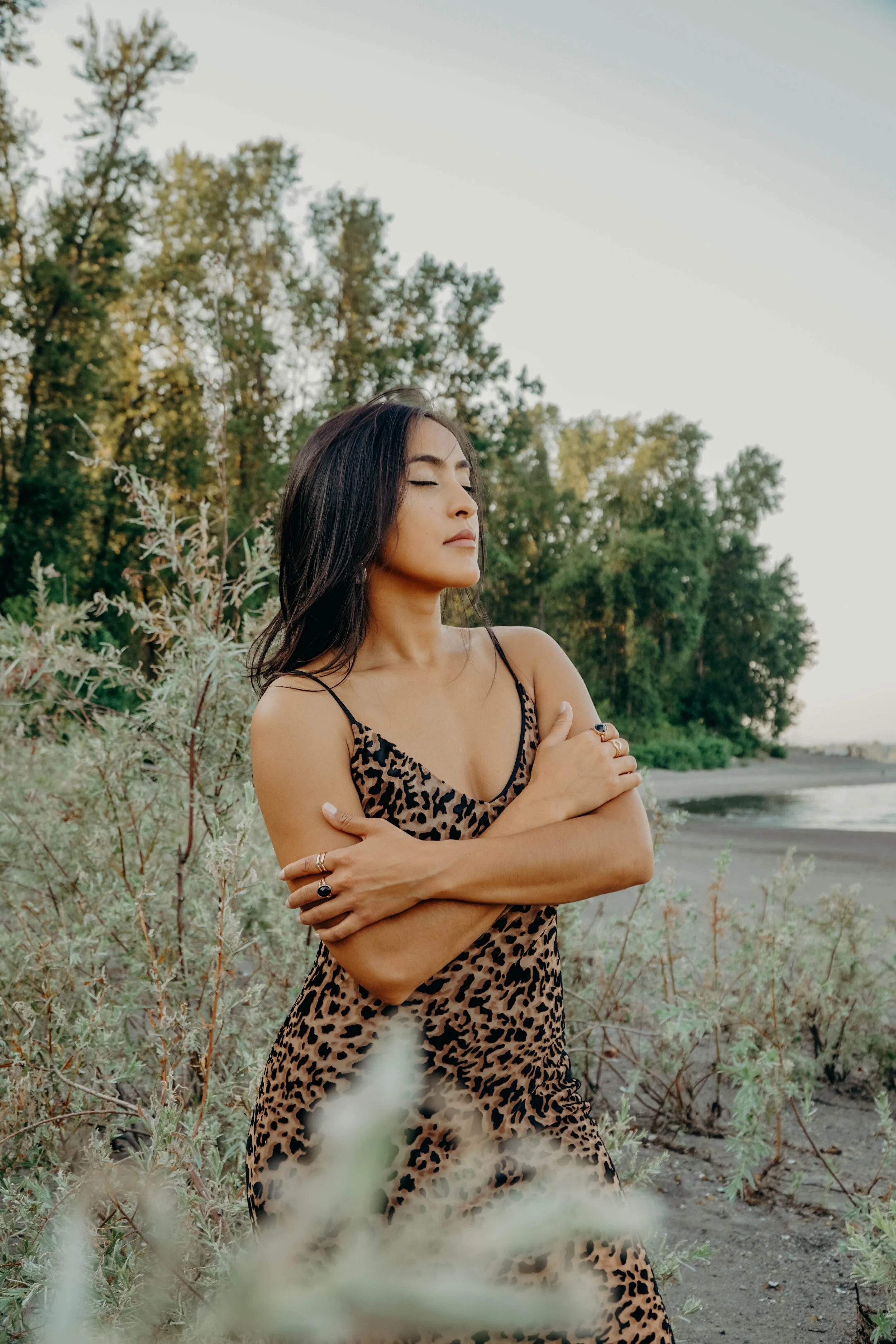 A woman with long dark hair and a leopard print dress stands with arms crossed, eyes closed, in a natural outdoor setting near a beach with trees and water in the background.