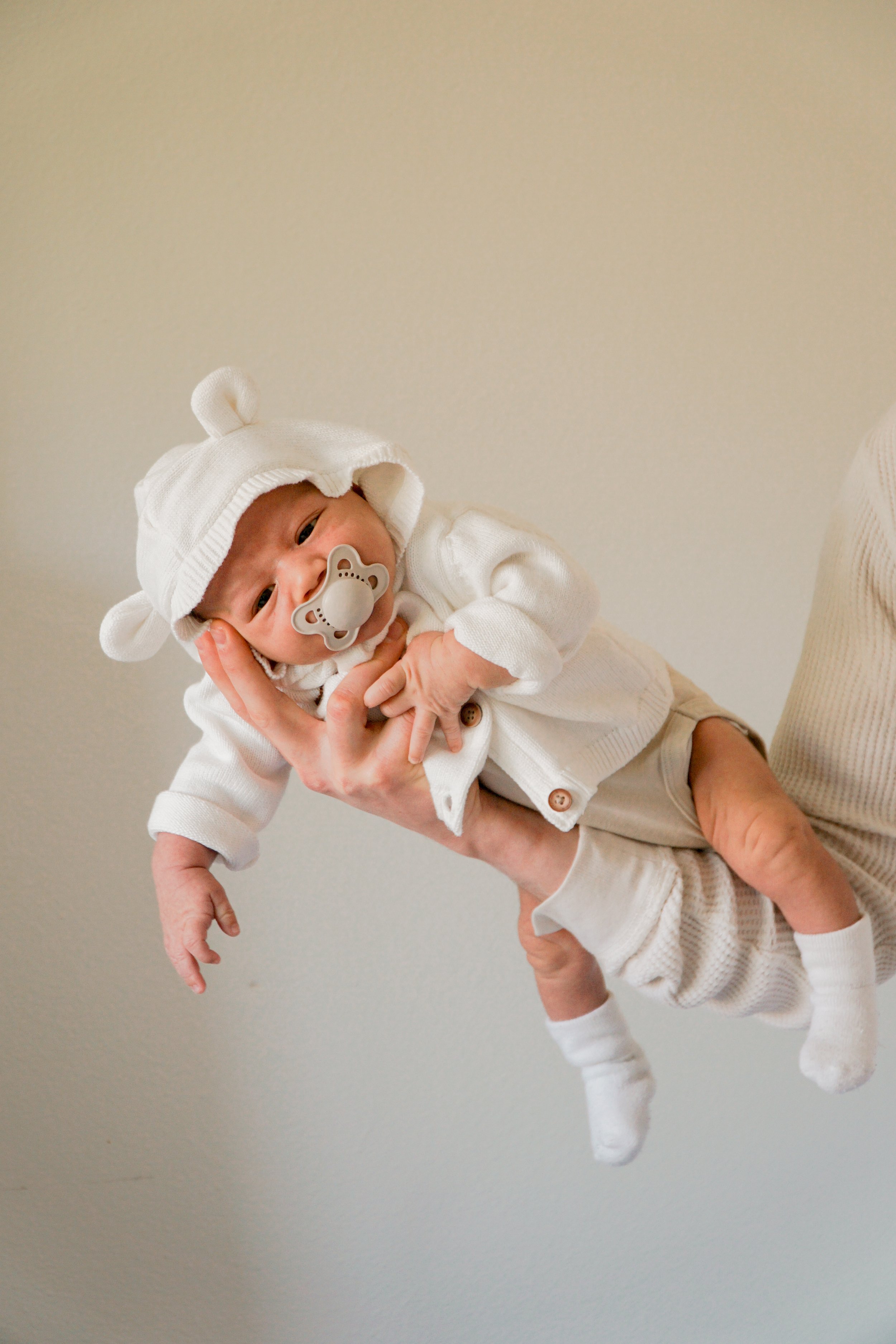 Baby dressed in white with a bear hat and pacifier, being held up by an adult, against a plain wall.