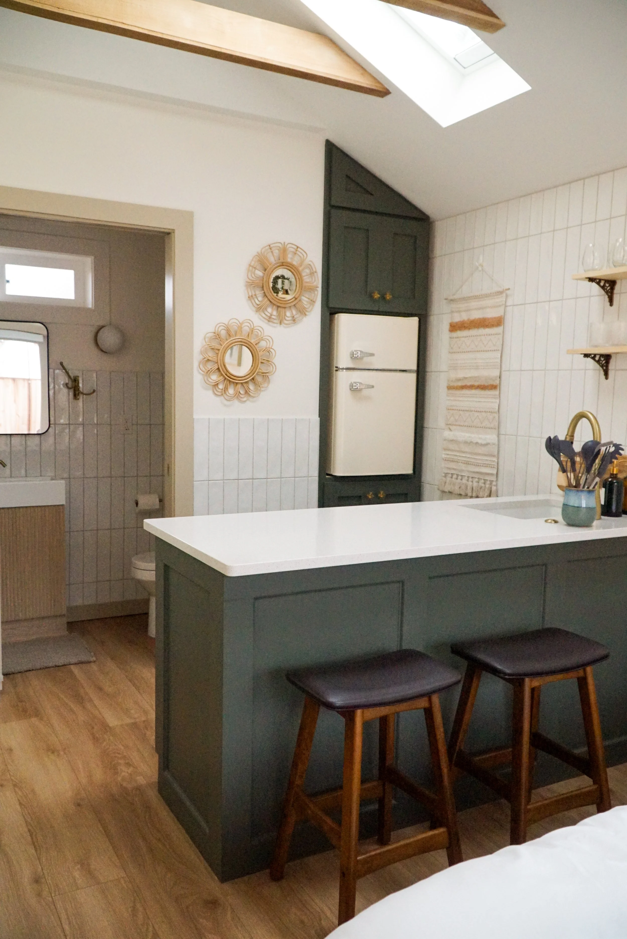 A cozy kitchen with a green island, wooden bar stools with black seats, a white countertop, and a wall with decorative woven mirrors and shelves.