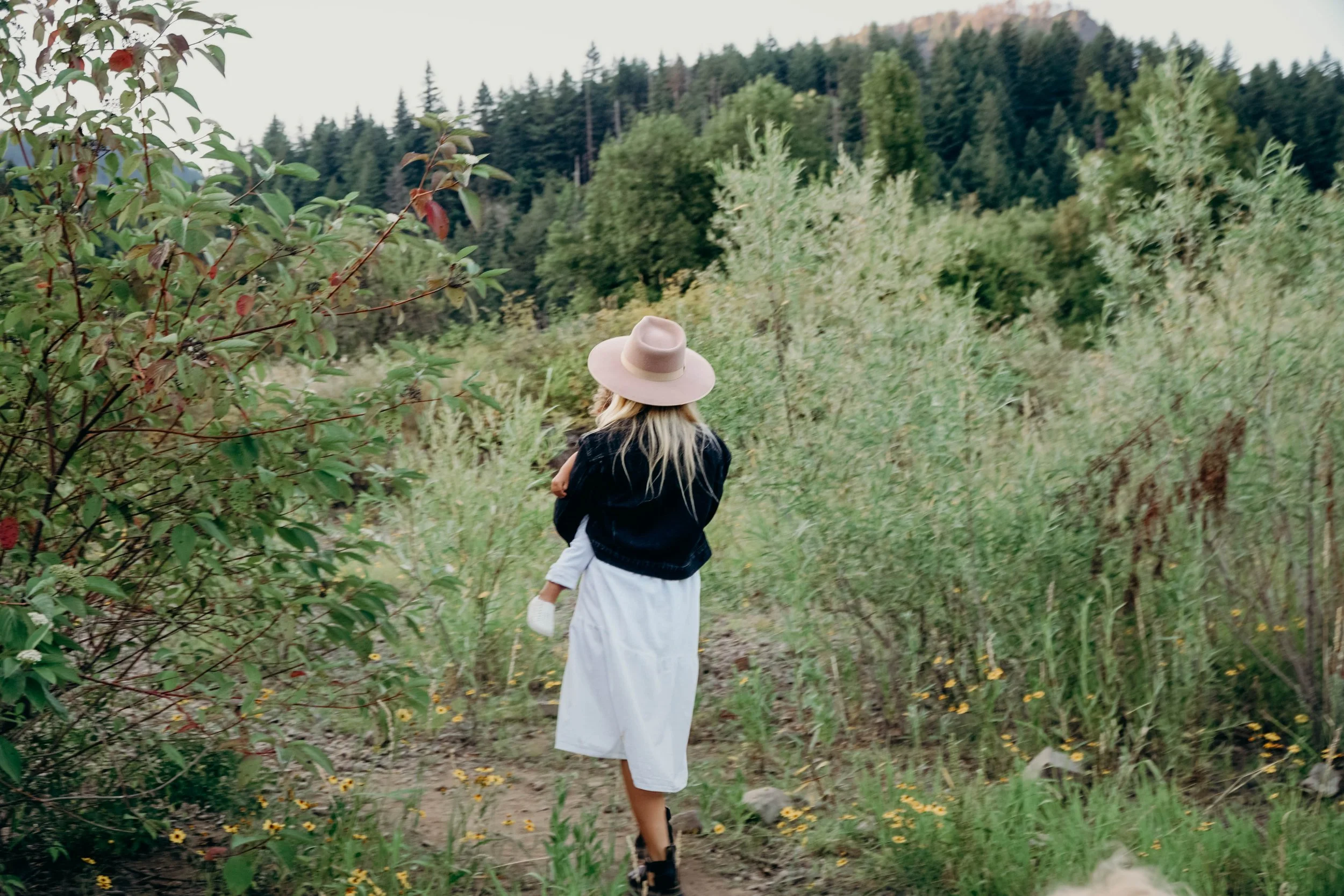 A woman walking on a trail through a lush green forest with hills and trees in the background, wearing a wide-brimmed hat, a black jacket, and a white dress.