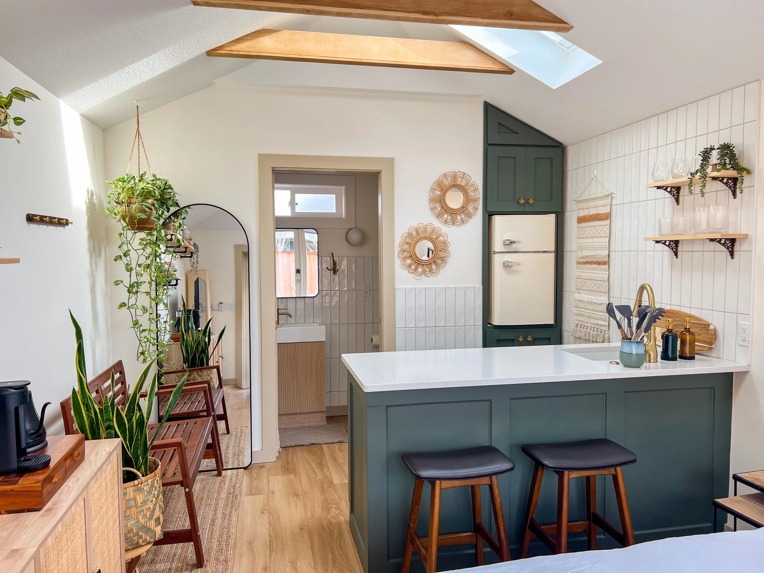 Cozy kitchen with a green island, white countertop, hanging plants, decorative mirrors, wooden shelves with glassware, and a small dining area with black stools.
