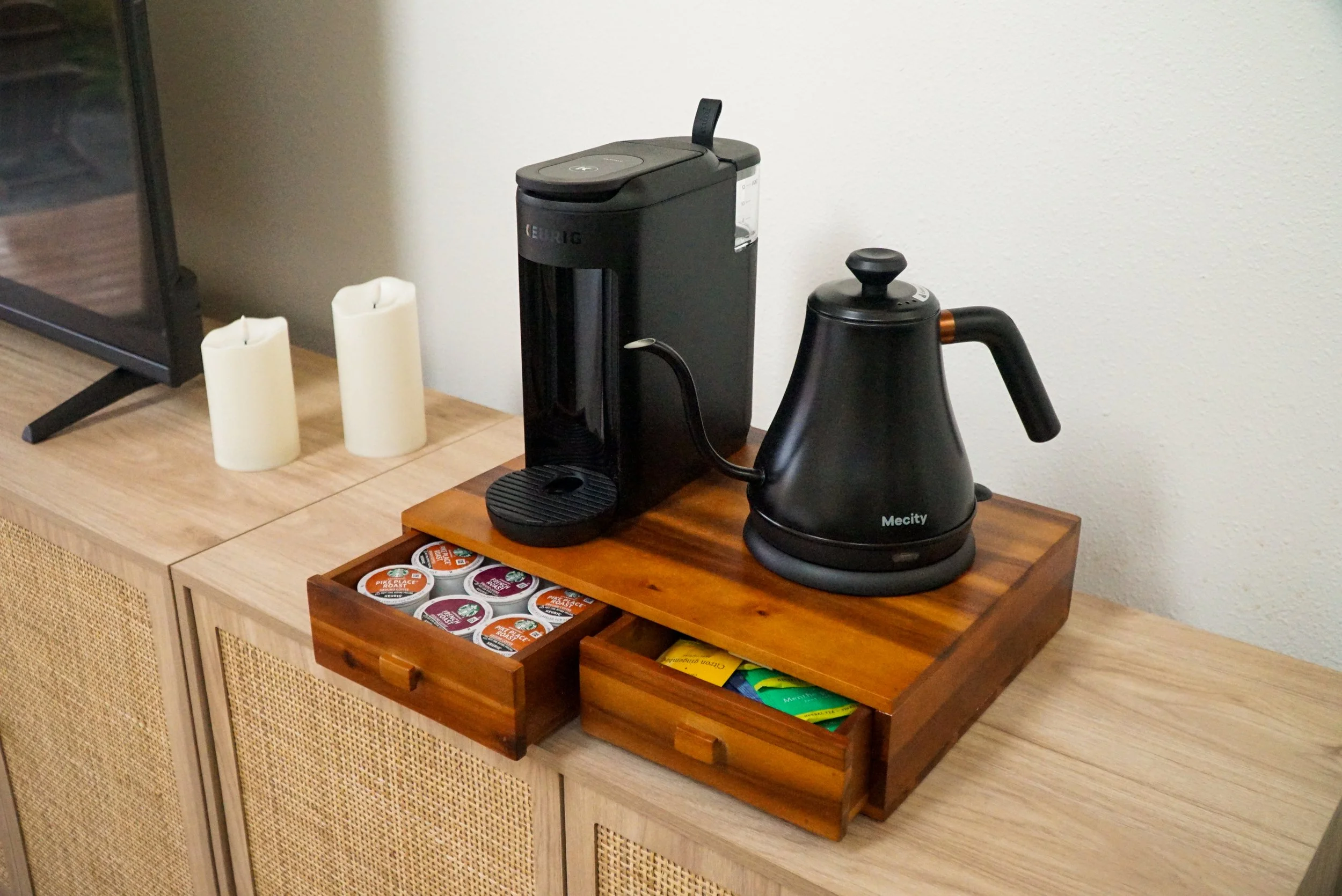 Coffee maker and electric kettle on a wooden tray, two white candles, and a TV on a wooden cabinet.