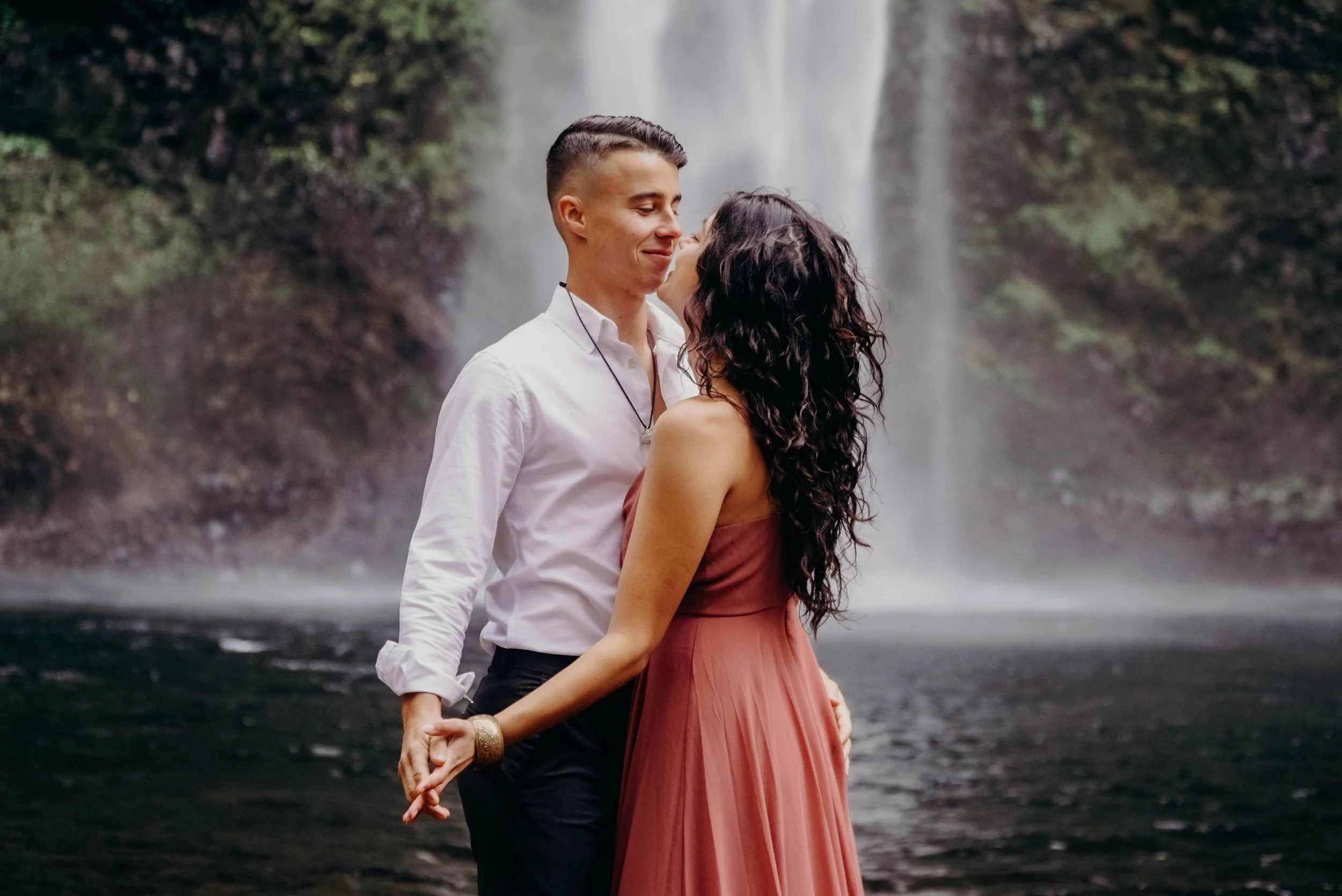 A couple stands close together near a waterfall, holding hands and smiling at each other. The man wears a white shirt, and the woman wears a pink dress with long curly hair.