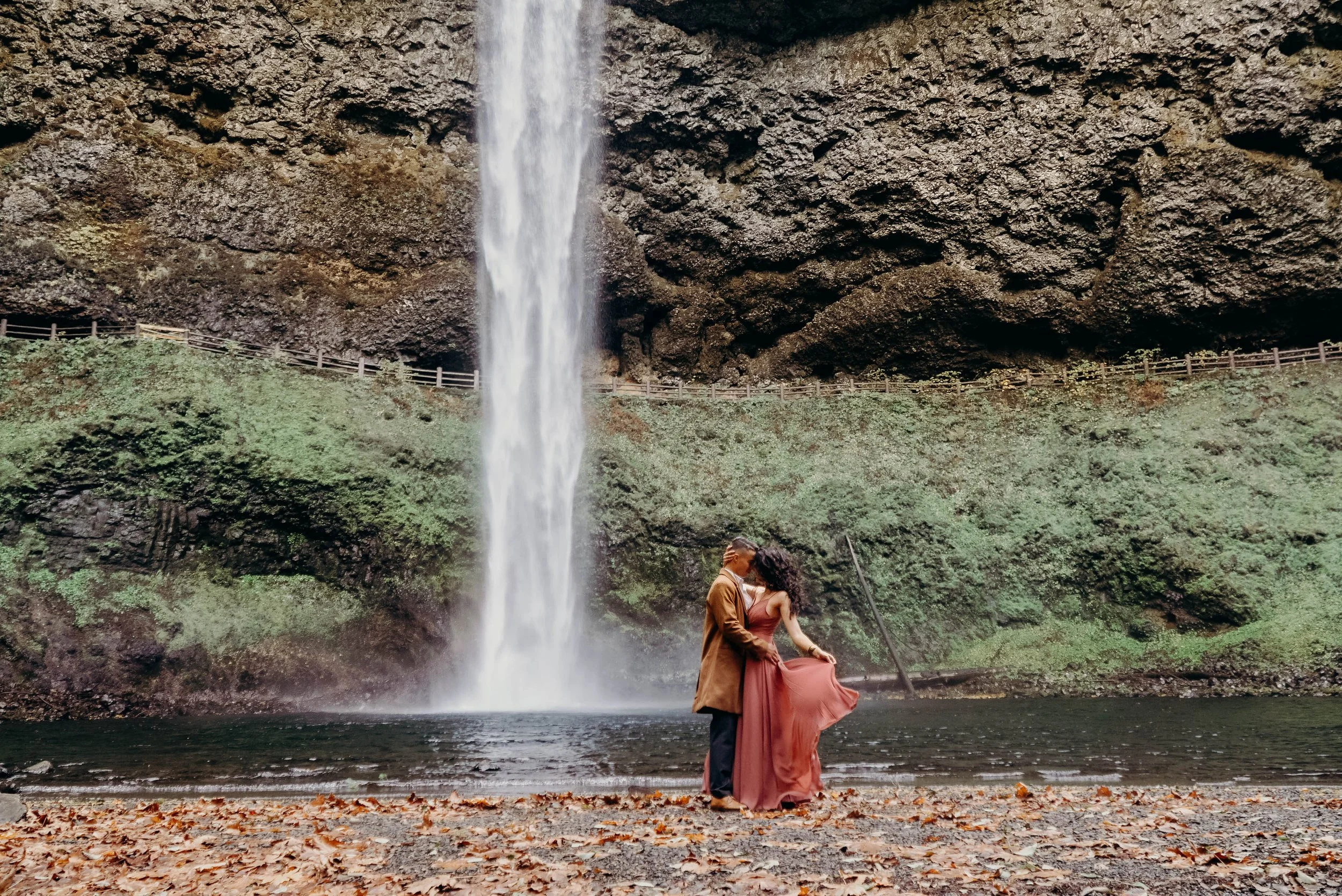A couple stands close together near a river, kissing, with a tall waterfall cascading in the background and a forested hillside.