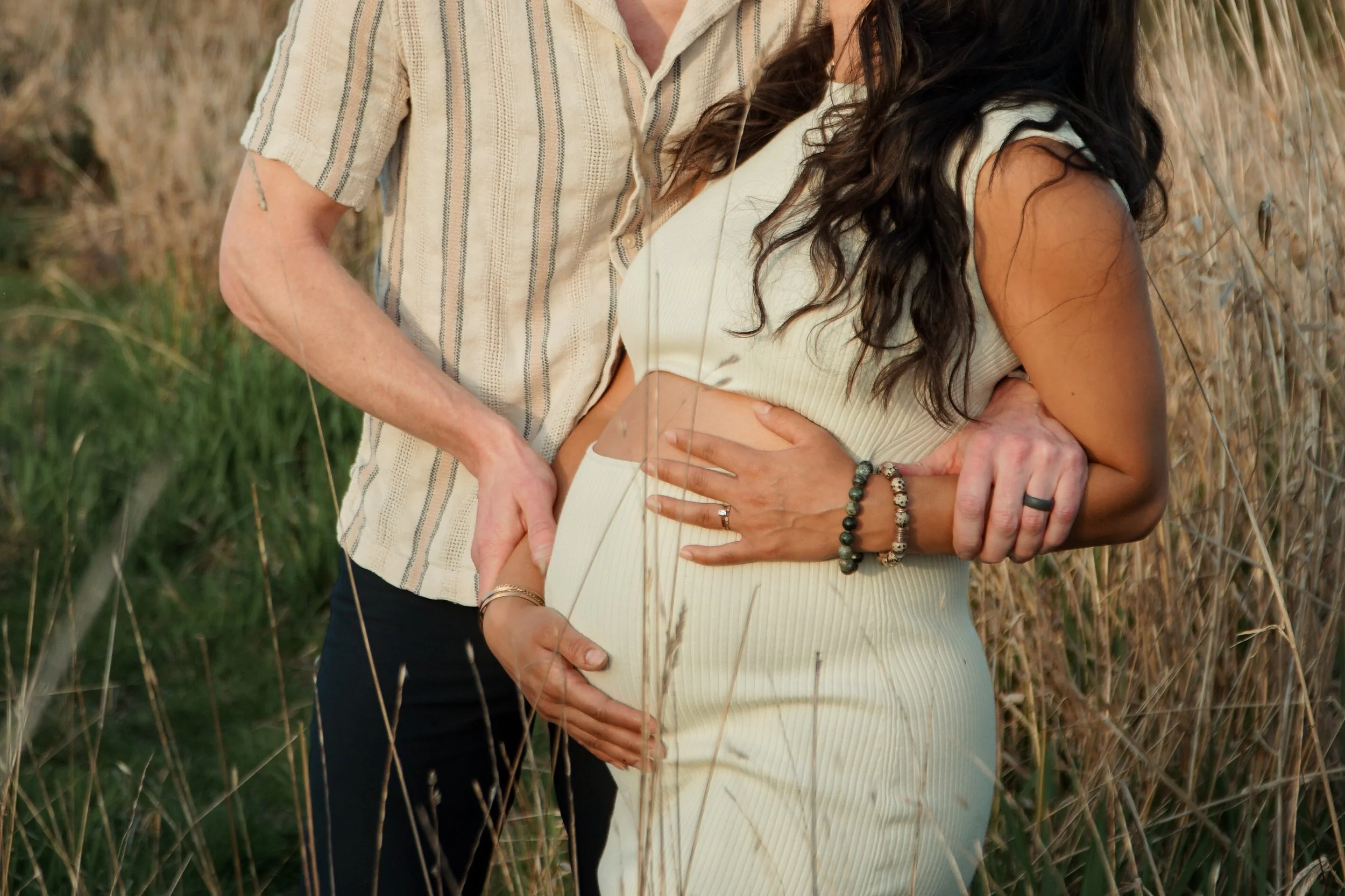 A pregnant woman and a man, possibly her partner, standing in a field of tall grass or wheat, with the man gently holding her belly from behind and both hands resting on her stomach. The woman is wearing a cream-colored, ribbed, sleeveless dress, and the man is wearing a light-colored, short-sleeved shirt with vertical stripes. The woman is accessorized with bracelets and rings, and has long dark hair.