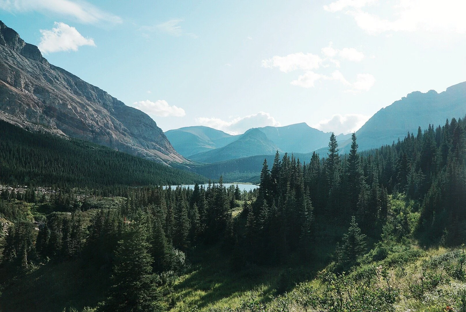 Scenic view of a mountain valley with dense evergreen forests, a lake, and mountain peaks under a partly cloudy sky.