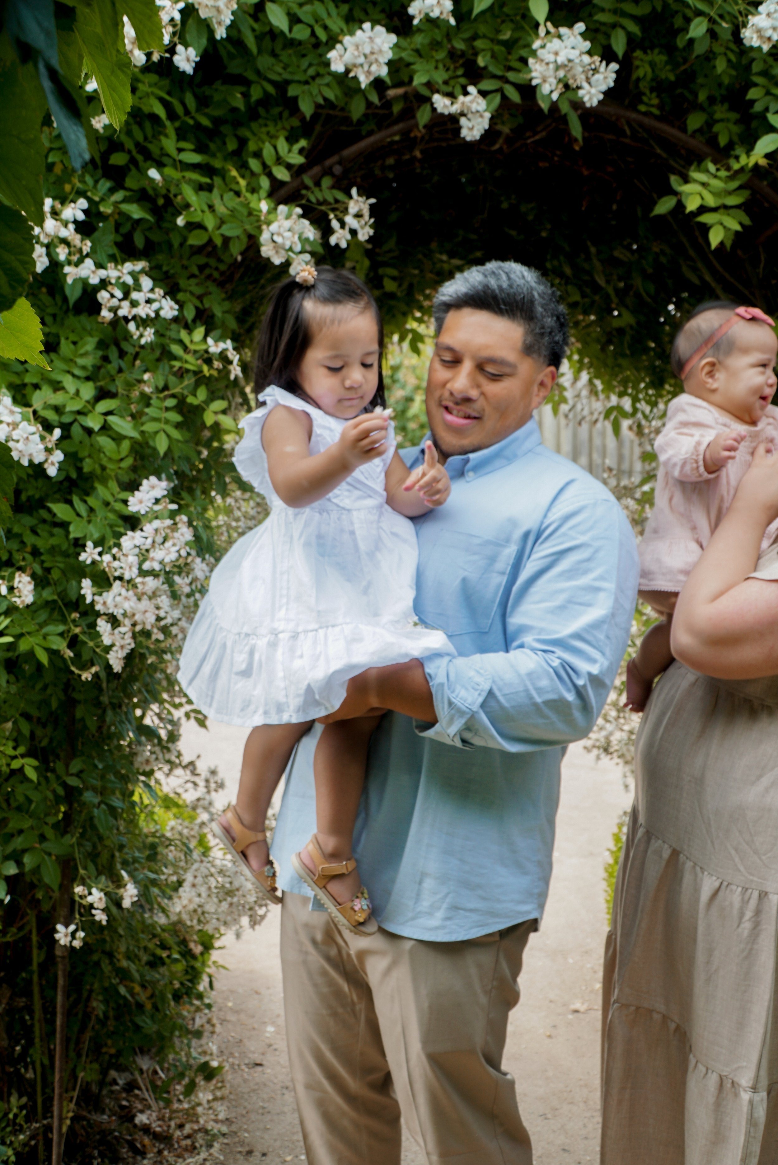 A man holding a young girl in a white dress under a floral archway, with a woman holding a baby nearby. The scene is outdoors among greenery and white flowers.